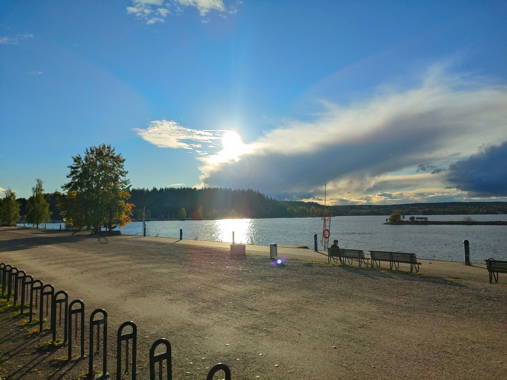 Lahti, Vesijärven satama alkuillasta. Rannan läheisyydessä näkyykö muutama penkki. Taivas on sininen, lähes pilvetön.  Aurinko alkaa vaipua alaspäin ja on juuri katoamassa pilven taakse vastarannalla. Vedessä näkyy heijastus, kuin  auringonsilta.