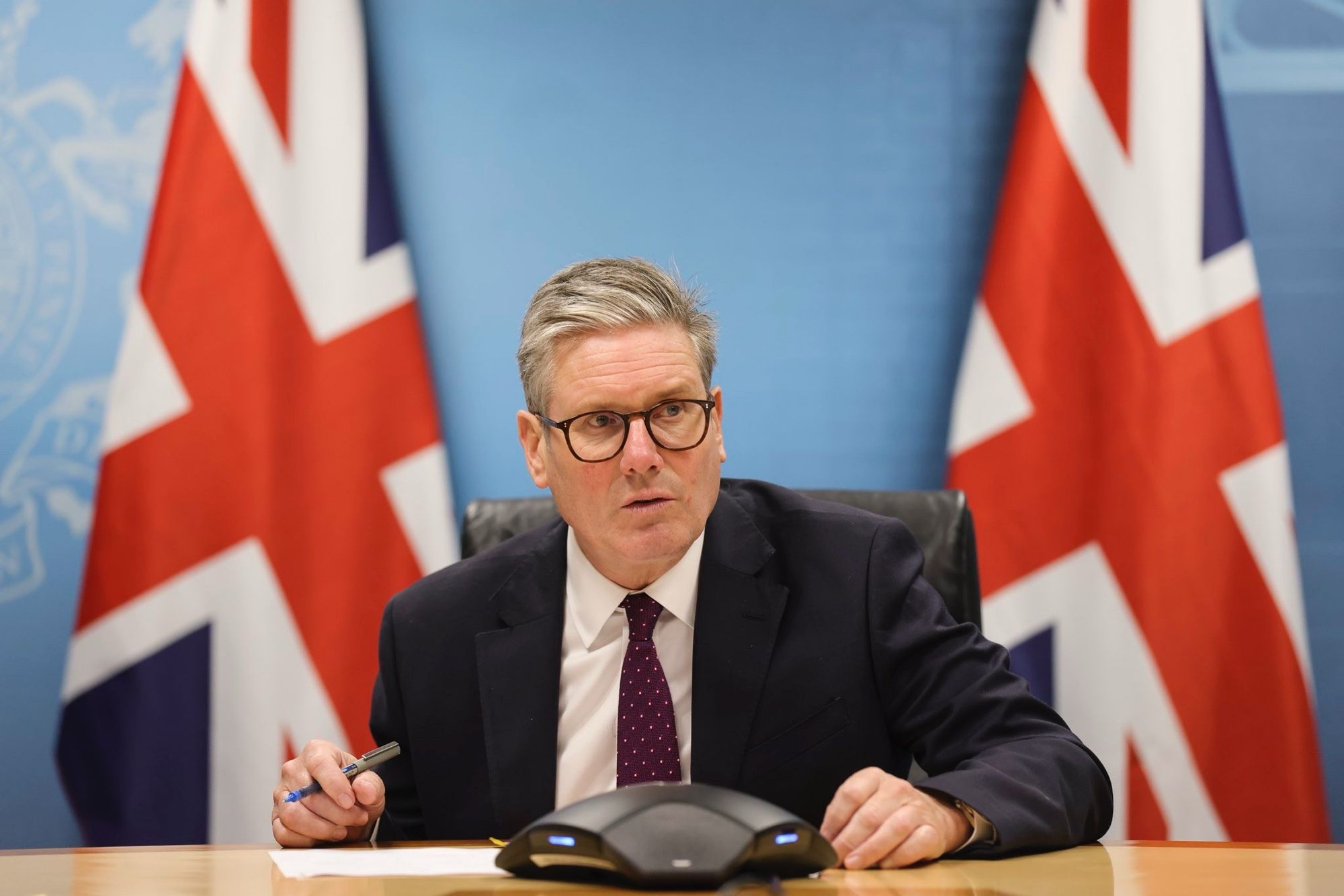 The right-wing Prime Minister of the UK, Keith Stormfront, pretending to be on a phone call with a comically inordinate number of union jacks behind him as if he’s doing a party political broadcast for the National Front