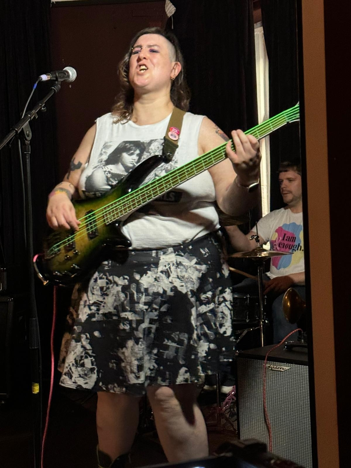 Pic of me (white nonbinary person) wearing white top and black and white skirt. I'm playing a green 5 string bass guitar. Behind me is the drummer of my band (white cis man)