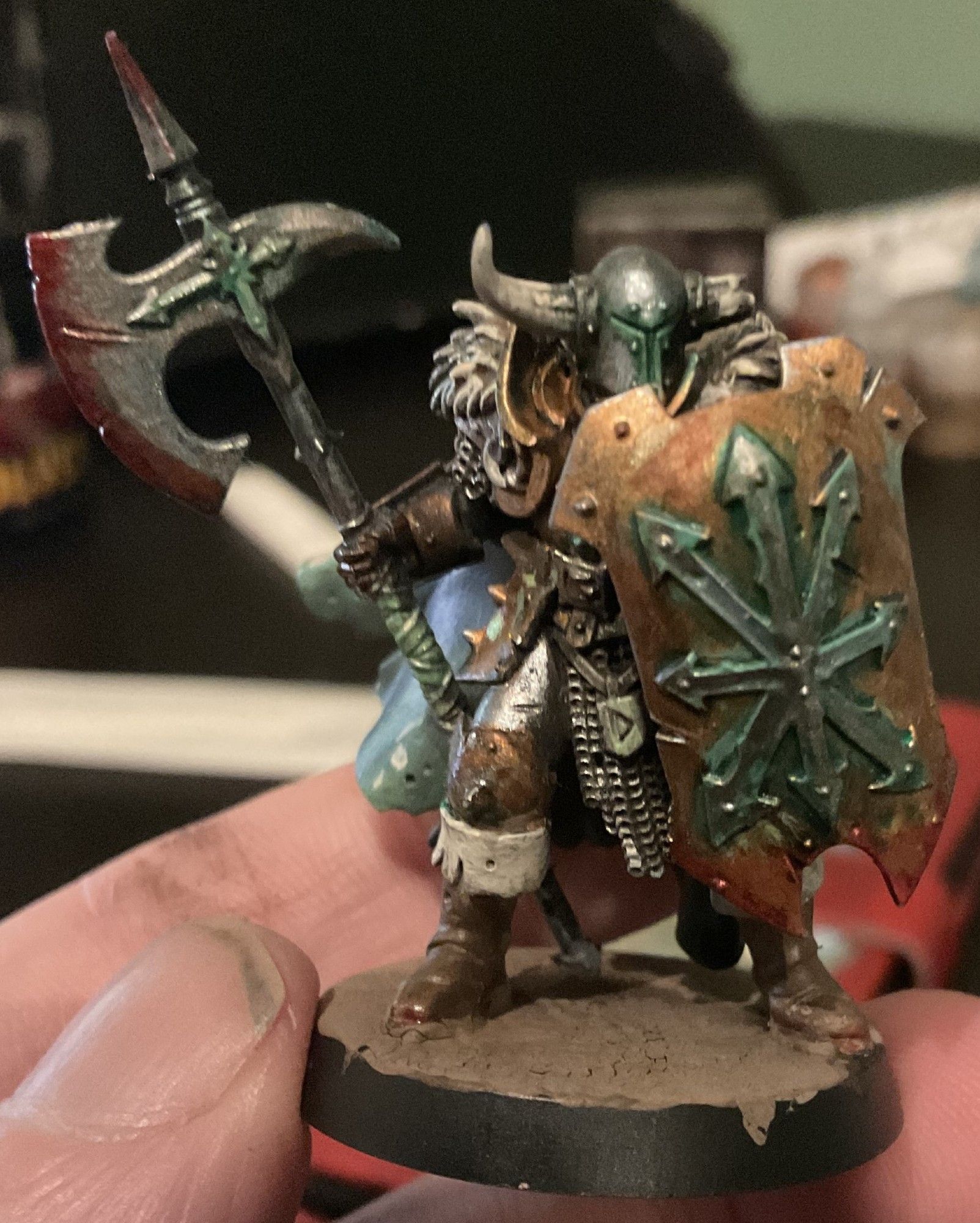 A Savage Slave to Darkness warhammer Age of sigmar Mini. It is holding a Shield with the Chaos symbol that has heavy oxidization around the symbol. They are wearing a horned helmet with one of the horns broken and is wearing brown leather boots and holding a halberd