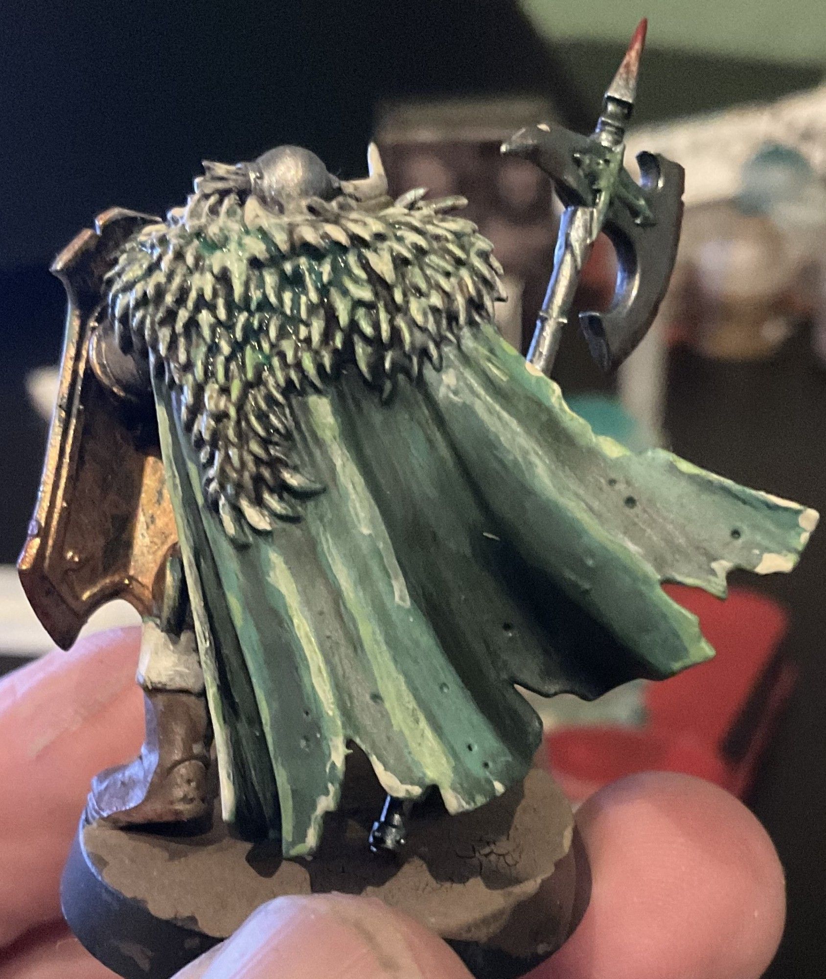 A Savage Slave to Darkness warhammer Age of sigmar Mini. It is holding a Shield l. They are wearing a horned helmet with one of the horns broken and is wearing brown leather boots and holding a halberd. This is a view from the back and he is earing a green cape with a fur pelt over his shoulders.