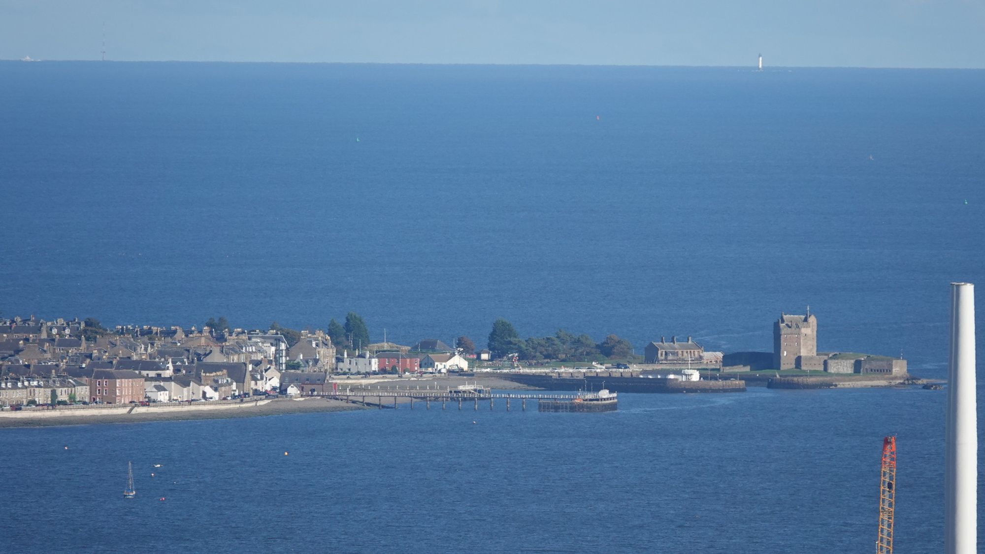 A zoom shot of a distance promontory with a castle amid deep blue water. The promontory extends from left to right with the castle at the right end. The left end is filled with houses. In the distance on the horizon a small white tower rises up: the Bell Rock lighthouse. A thin band of pale blue sky is above the water. A white wind turbine tower stands in the docks at the bottom right of the image with the top of a red crane beside it.