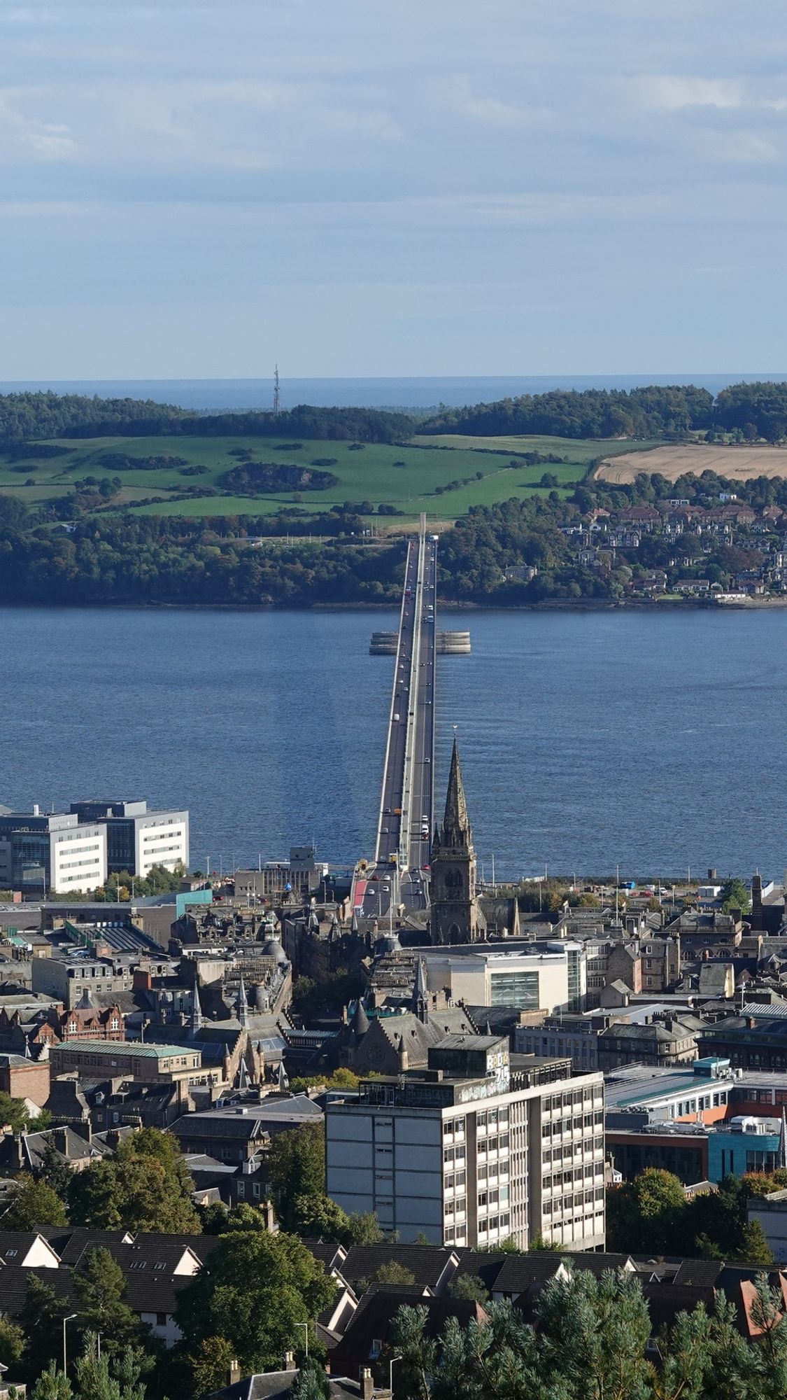 A portrait view looking down from the top of a hill on a road bridge crossing a wide blue river from the top of a hill. The foreground bottom third of the image has buildings and a spire; on the far side there are houses on the right of the bridge and green fields to the left and into the distance. Above the fields is pale blue cloudy sky.