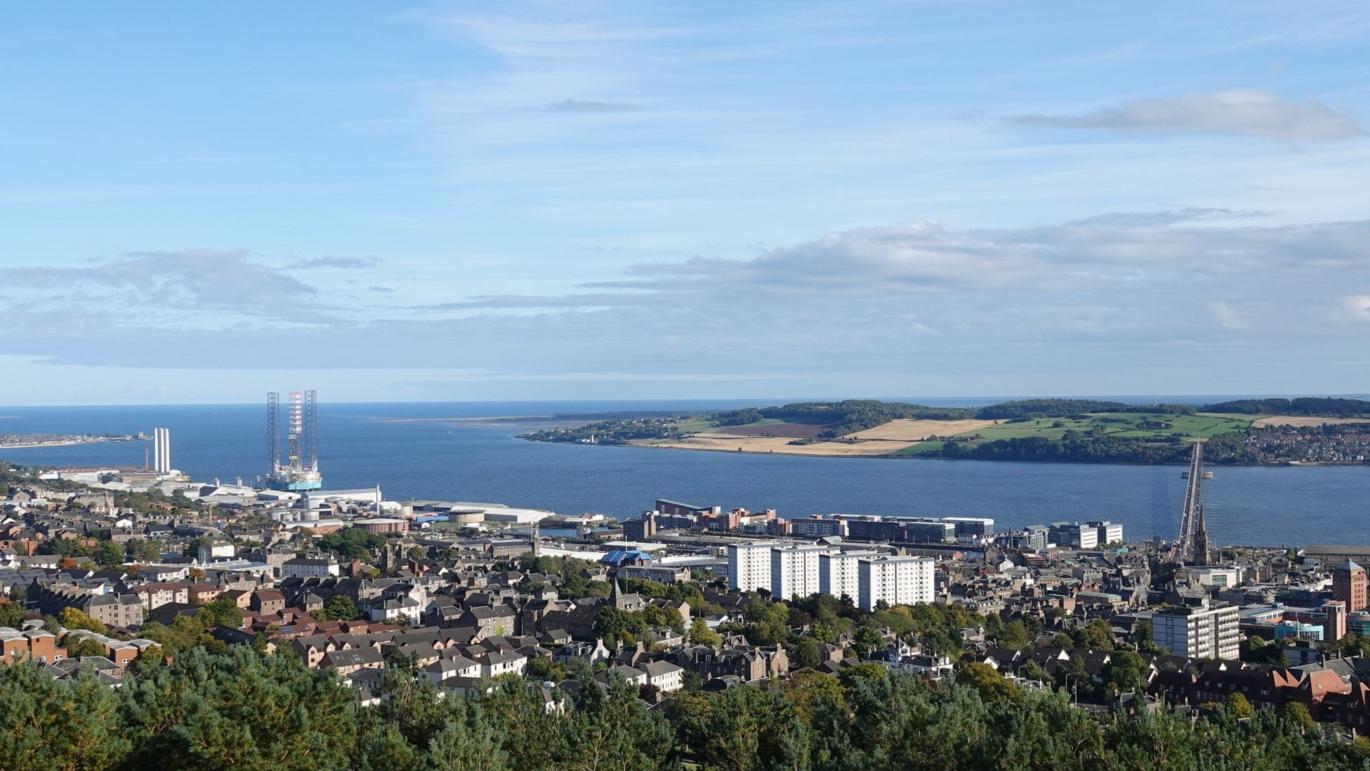 A wide scenic view of the estuary of the River Tay from the top of a hill. The Dundee fills the bottom third of the picture with the blue river above from left to right, and green and yellow fields of Fife in the distance to the middle. The top half of the picture is pale blue sky with grey and white streaky clouds. On the left in the distance are wind turbine towers and an oil rig. On the right a road bridge crosses the river directly away from the camera.