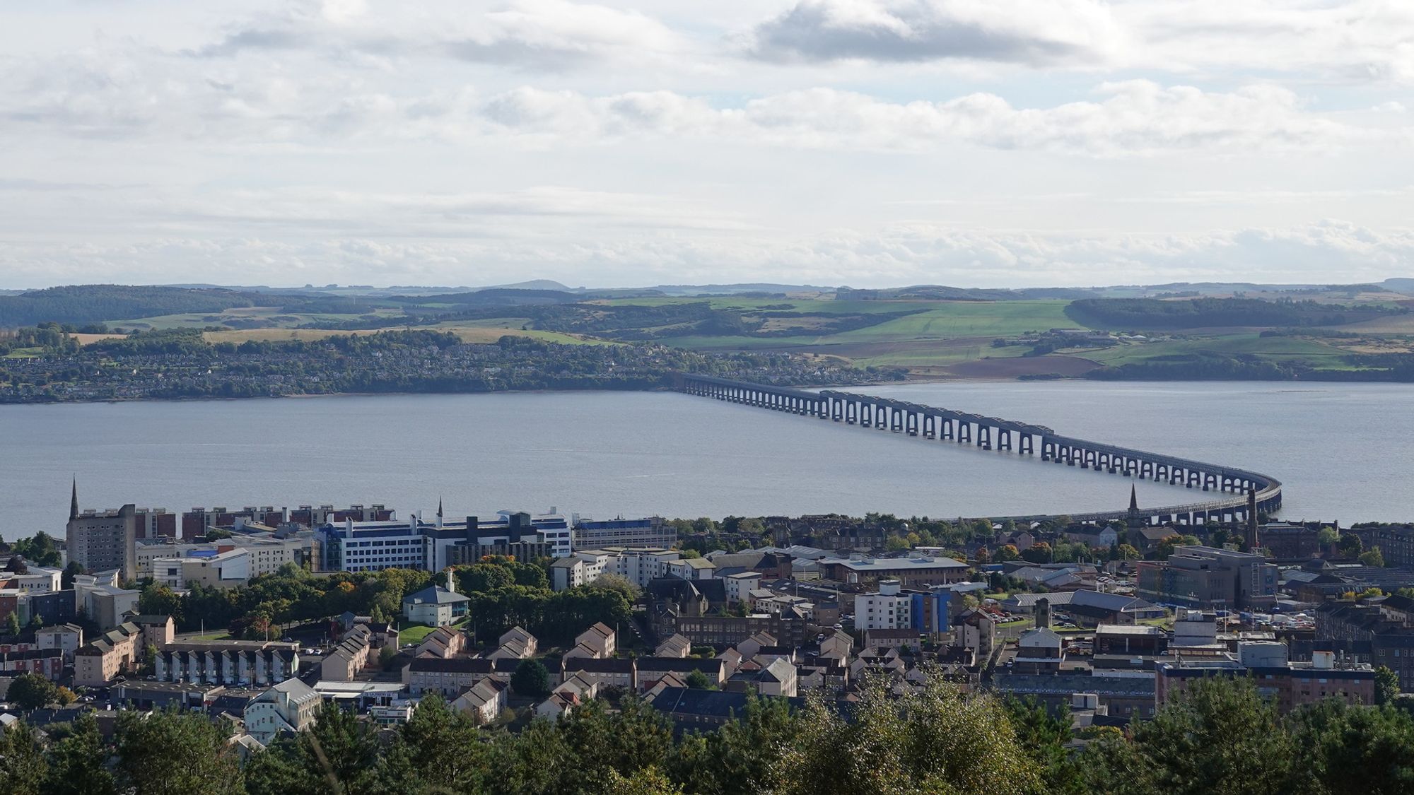 A wide scenic view of the River Tay and the Tay Rail bridge viewed from the top of a hill. The Dundee fills the bottom third of the picture with the pale silvery river above it from left to right. The bridge emerges roughly parallel to the bank towards the right of the picture then curves around and crosses the river diagonally. On the dar side is a long low hill with green and yellow fields and woods. The top of the picture is pale blue sky with grey and white streaky clouds.