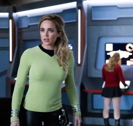 Captain Sara Lance of the Waverider (Caity Lotz, Legends of Tomorrow) stands on a bridge reminiscent of the USS Enterprise, and wearing a pale lime green uniform shirt with silver striped cuffs, black leather pants, and a phaser-style pistol on her hip.