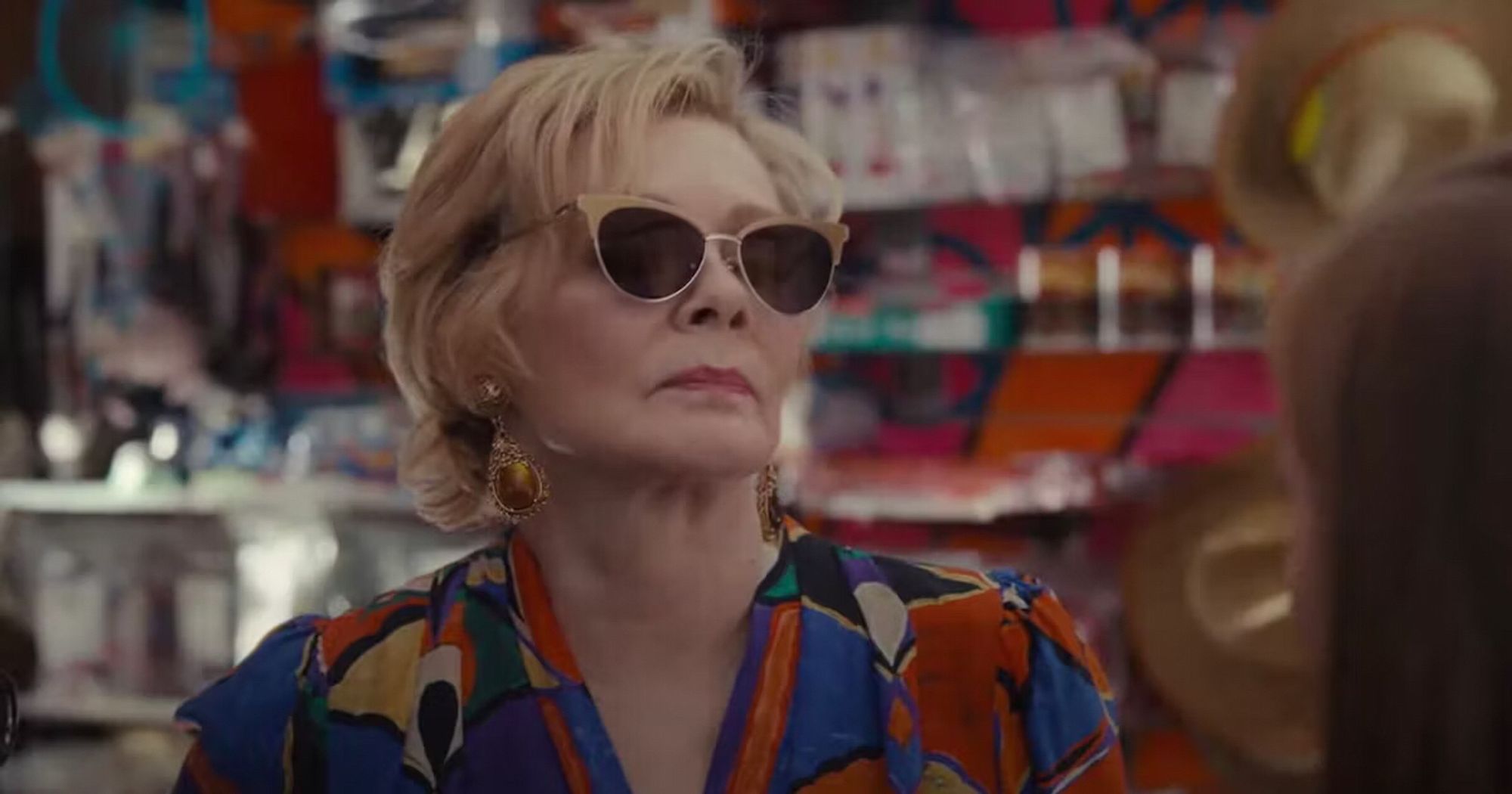 Deborah Vance (Jean Smart, Hacks) wearing a colorful blouse and gold cateye sunglasses in a roadside convenience store.