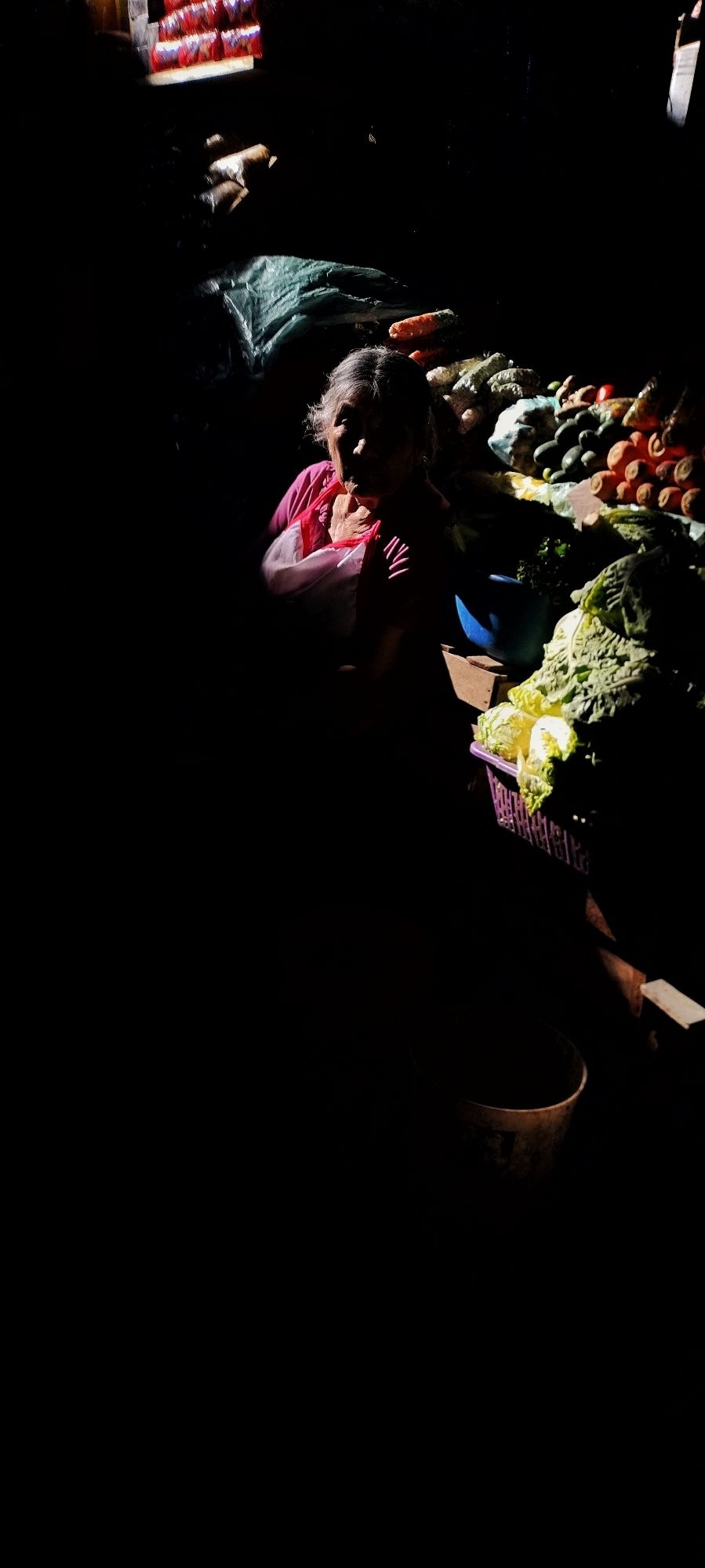 A senior woman vendor sits in a dimly lit market stall in Huánuco, Peru. Partially illuminated, she is surrounded by a display of fresh vegetables, including prominently placed carrots and lettuce. The dramatic contrast between light and shadow emphasizes her quiet presence amid the bustling market.