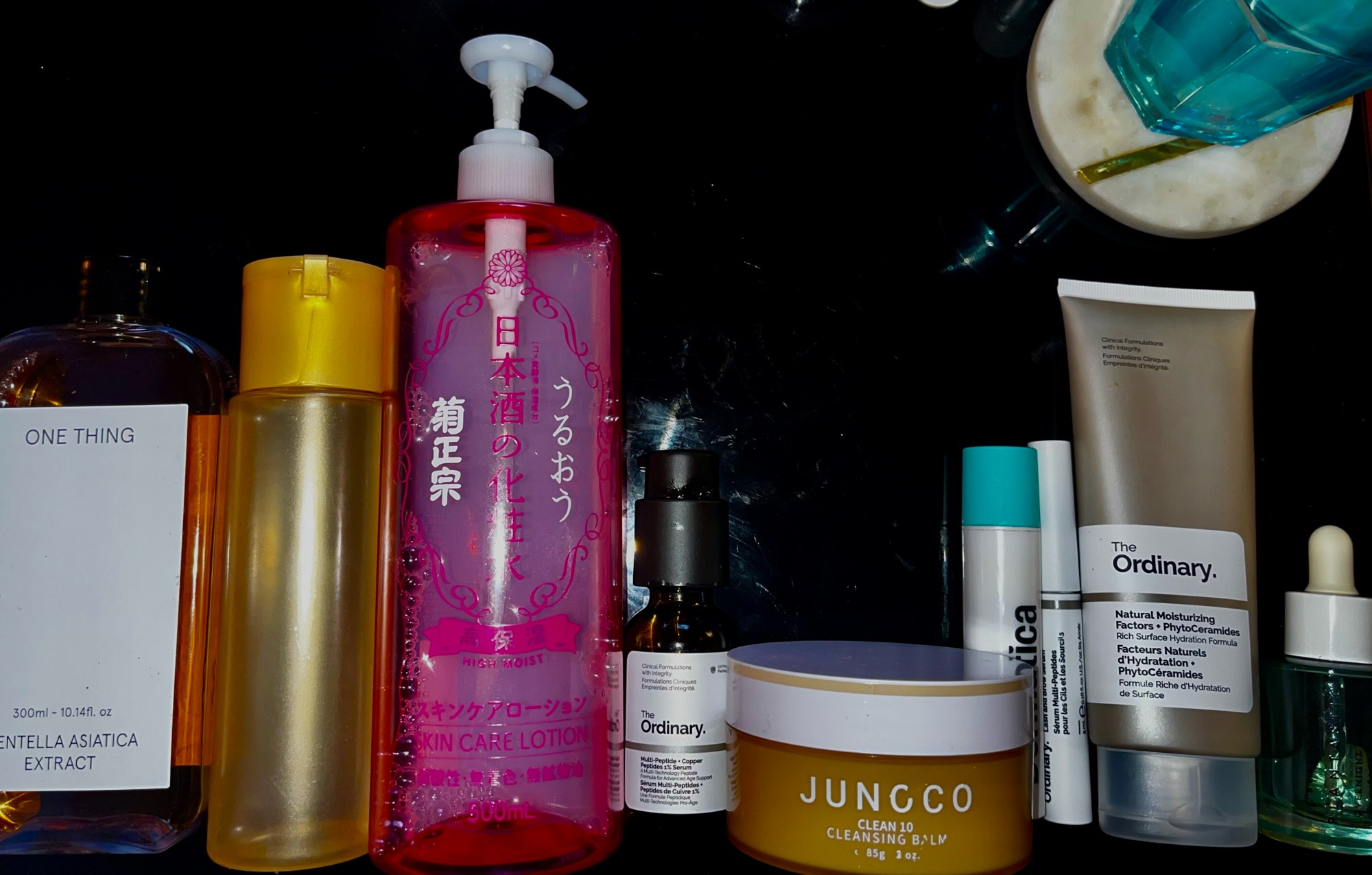 Image: a lineup of skincare products I use in my evening routine.
Clear bottle One Thing Centella Asiatica toner, yellow bottle without label Hadalabo Gyokujun Premium Lotion, large pink bottle Kikumasamune High Moist Lotion, brown bottle The Ordinary Multi Peptides + Copper Serum, orange jar Junc Co Cleansing Balm, white tube Dermatica, white tube, white tube The Ordinary Multi-Peptide Brow Serum, grey tube The Ordinary Natural And Factors + Phyto-Ceramides, clear bottle Beauty Pie Plantastic Oil.