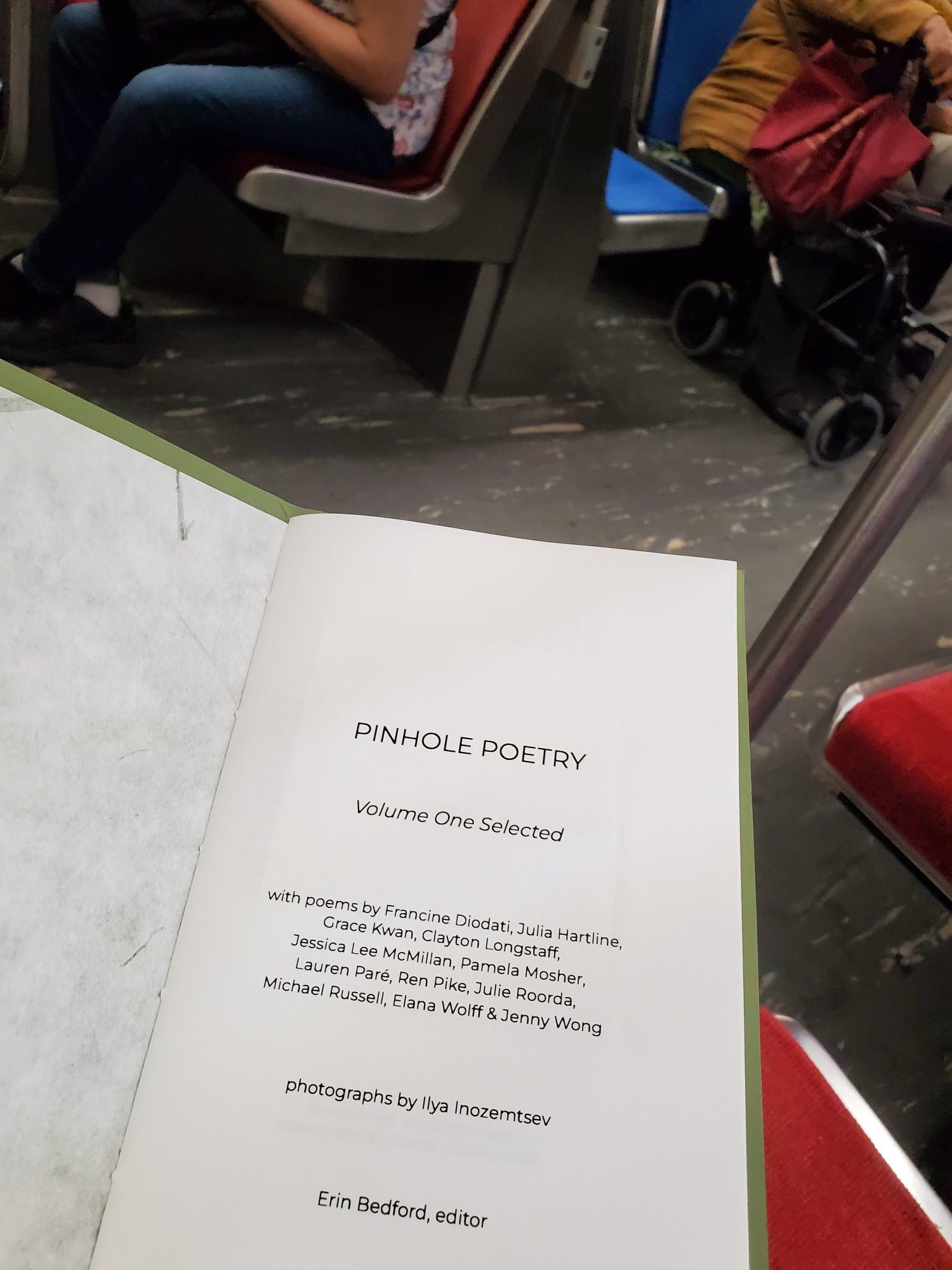 Reading the poetry collection/chapbook Pinhole Poetry Volume One Selected while riding the subway, with subway seats and passengers visible in the background