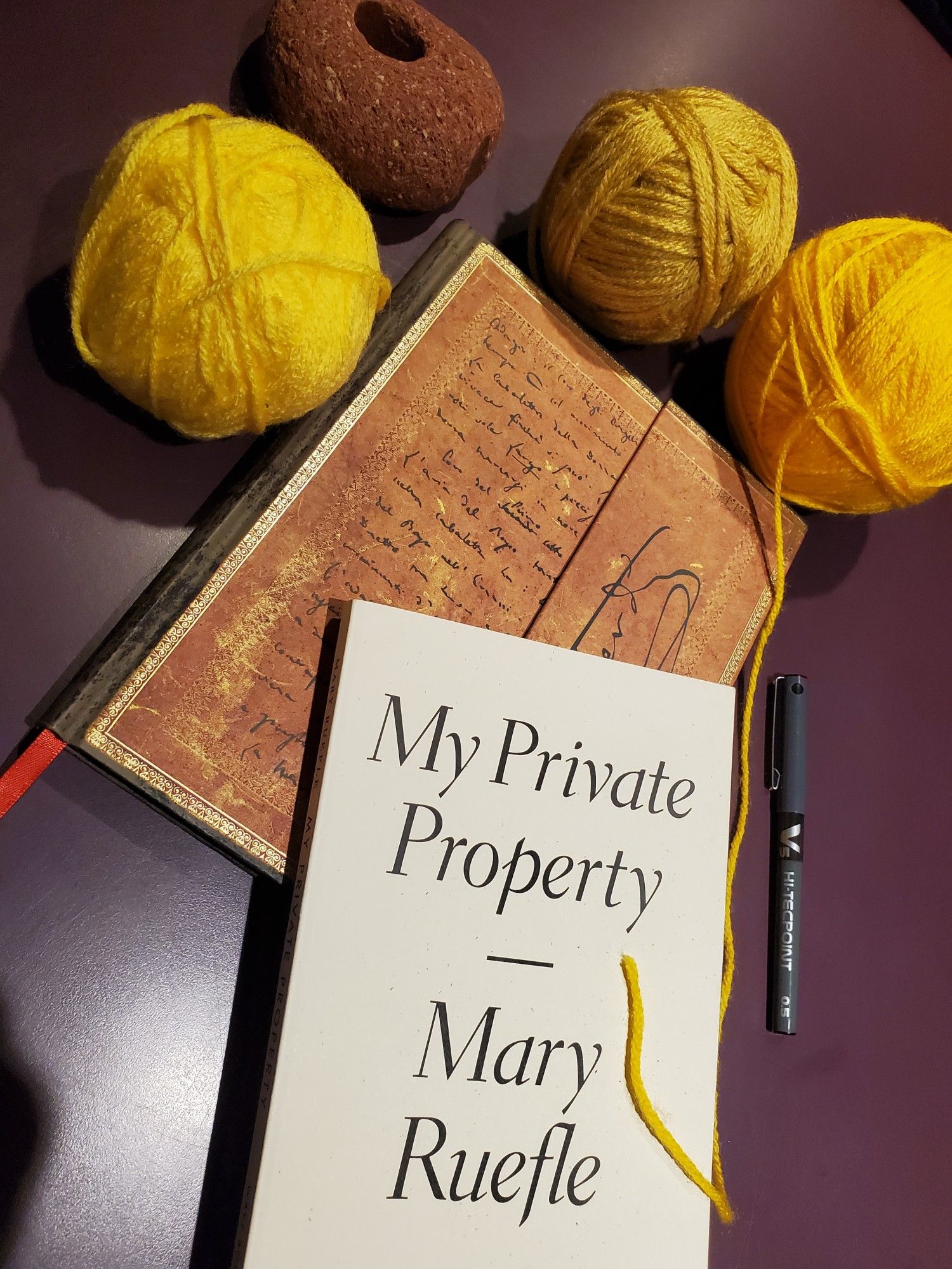 Poetry collection My Private Property by Mary Ruefle (Wave Books) sits atop a notebook with an ornate gold cover on a purple desktop next to a piece of red brick and balls of yellow wool