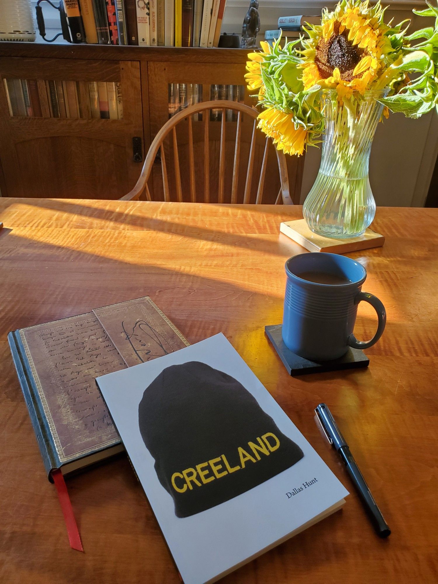 Poetry collection Creeland by Dallas Hunt (Nightwood Editions) sits on a sun-streaked dining room table with a notebook with an ornate cover, a black pen, coffee in a blue cup and a vase of slightly wilted sunflowers