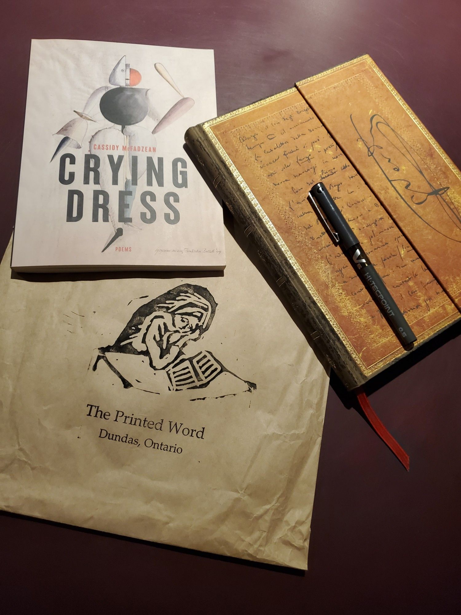 Poetry collection Crying Dress by Cassidy McFadzean (House of Anansi Press) sits on a purple desktop with a notebook with an ornate gold cover, a black pen and a paper bag from The Printed Word bookshop