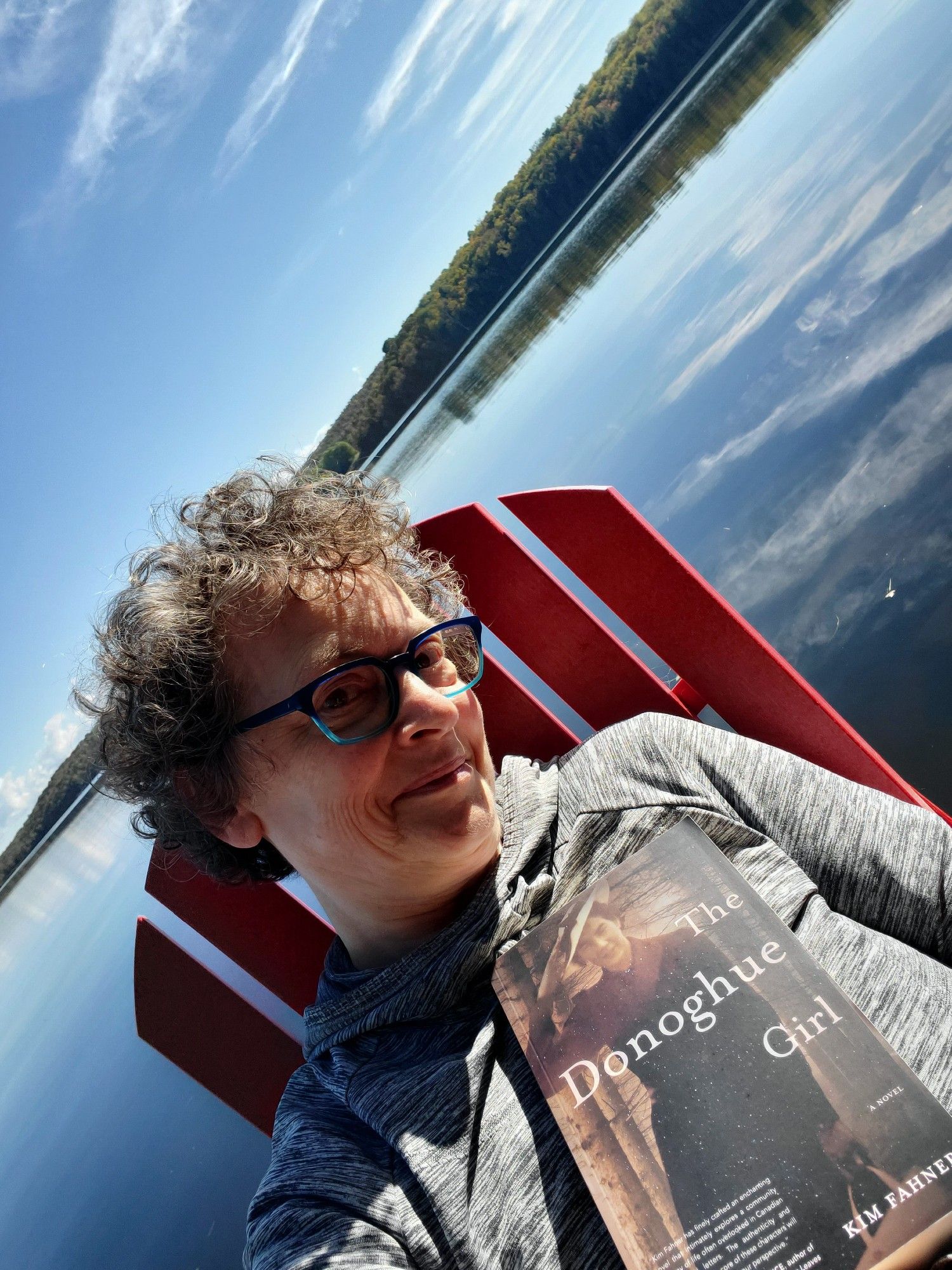 Me, sitting in a red chair on our dock on Big Doe Lake, holding the book The Donoghue Girl by Kim Fahner (Latitude 46 Publishing). The blue sky sports wispy clouds, and the lake and far shore shine in the morning sunlight.