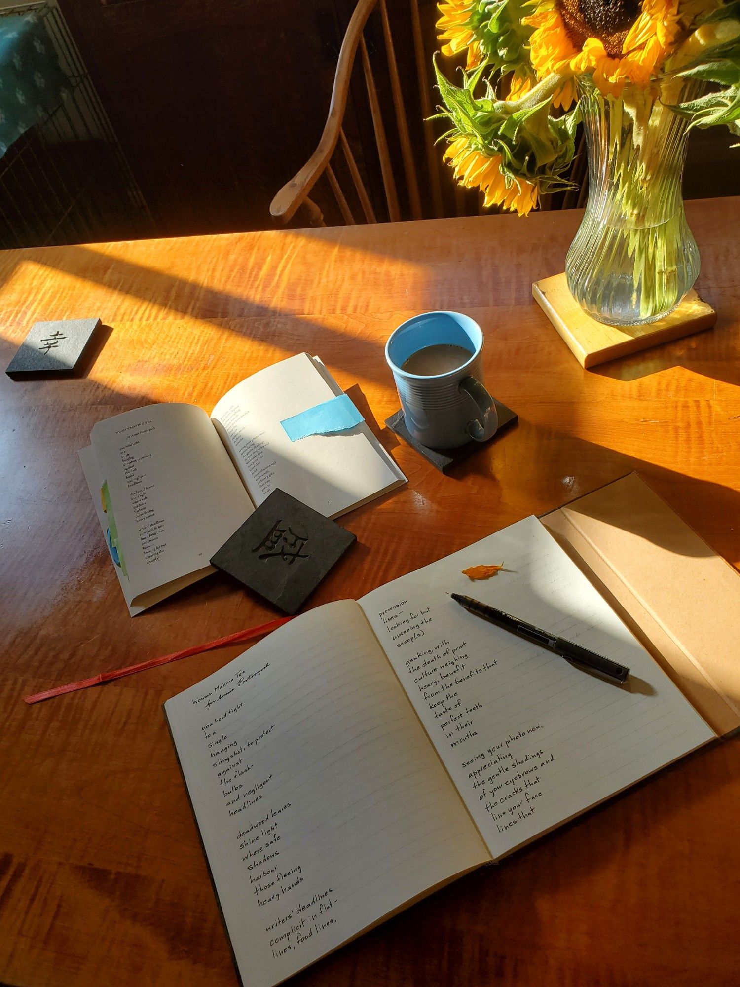 Handwritten transcription of the poem "Woman Making Tea" from the poetry collection Creeland by Dallas Hunt, with the poetry collection held open with a slate coaster, and an uncapped black pen and a sunflower petal resting on the notebook page