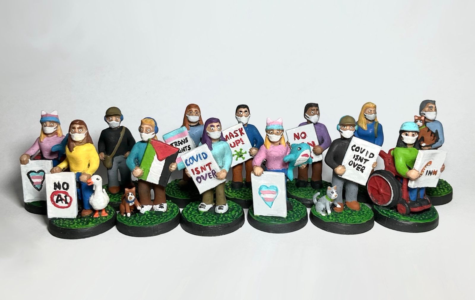 A group of 35mm 3D printed models designed by me in Nomad Sculpt and painted by me using acrylic paints. They wear a variety of colourful clothing and hats. All wear FFP3 masks. Some carry pets and mascots. Some are in wheelchairs and some carry placards with slogans like 'No AI', 'Covid isn't over', 'Trans Rights' etc