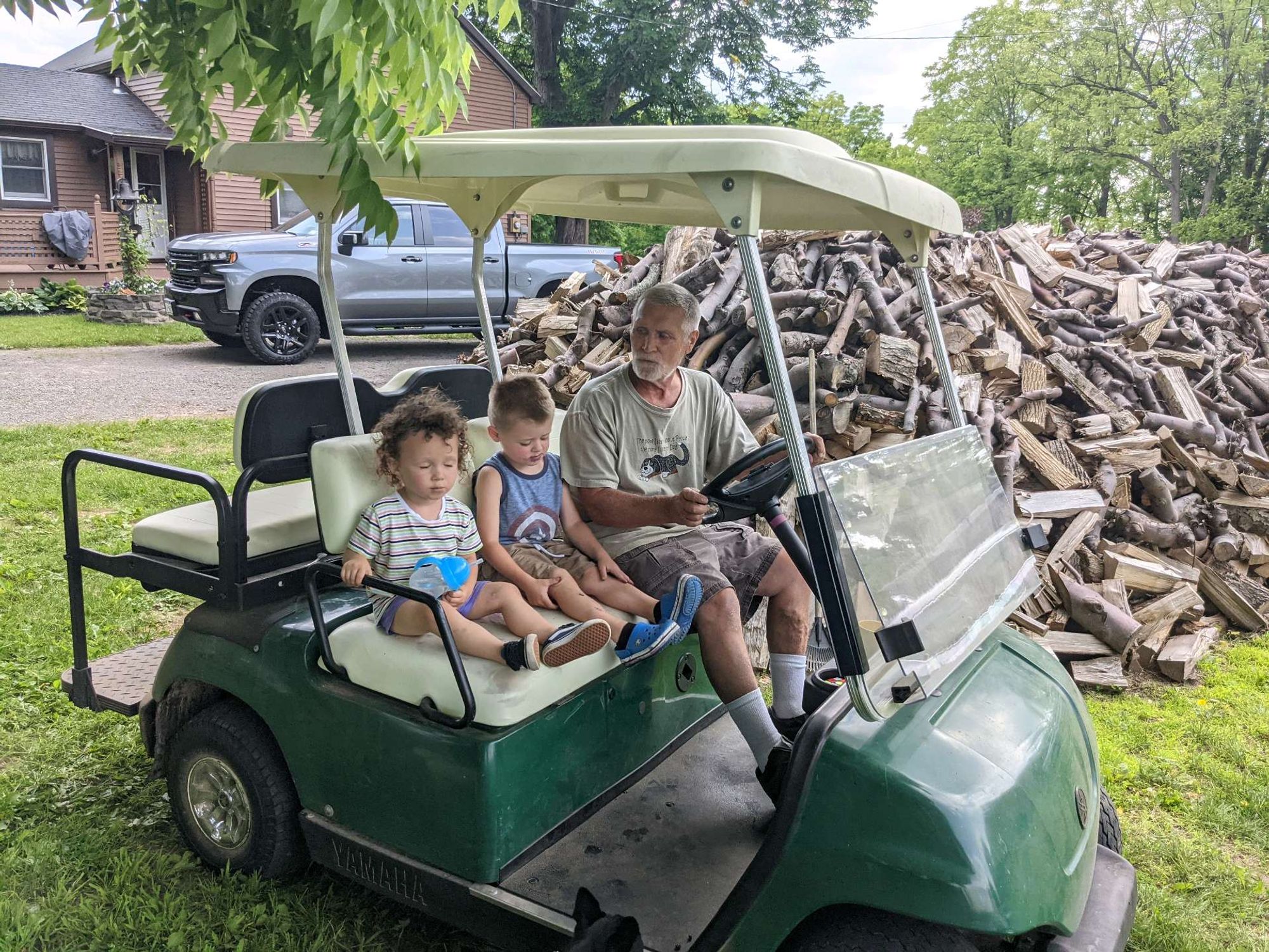 Man on a golf cart with two toddlers. The golf cart is in front of a large pile of wood. There's a house in the background 