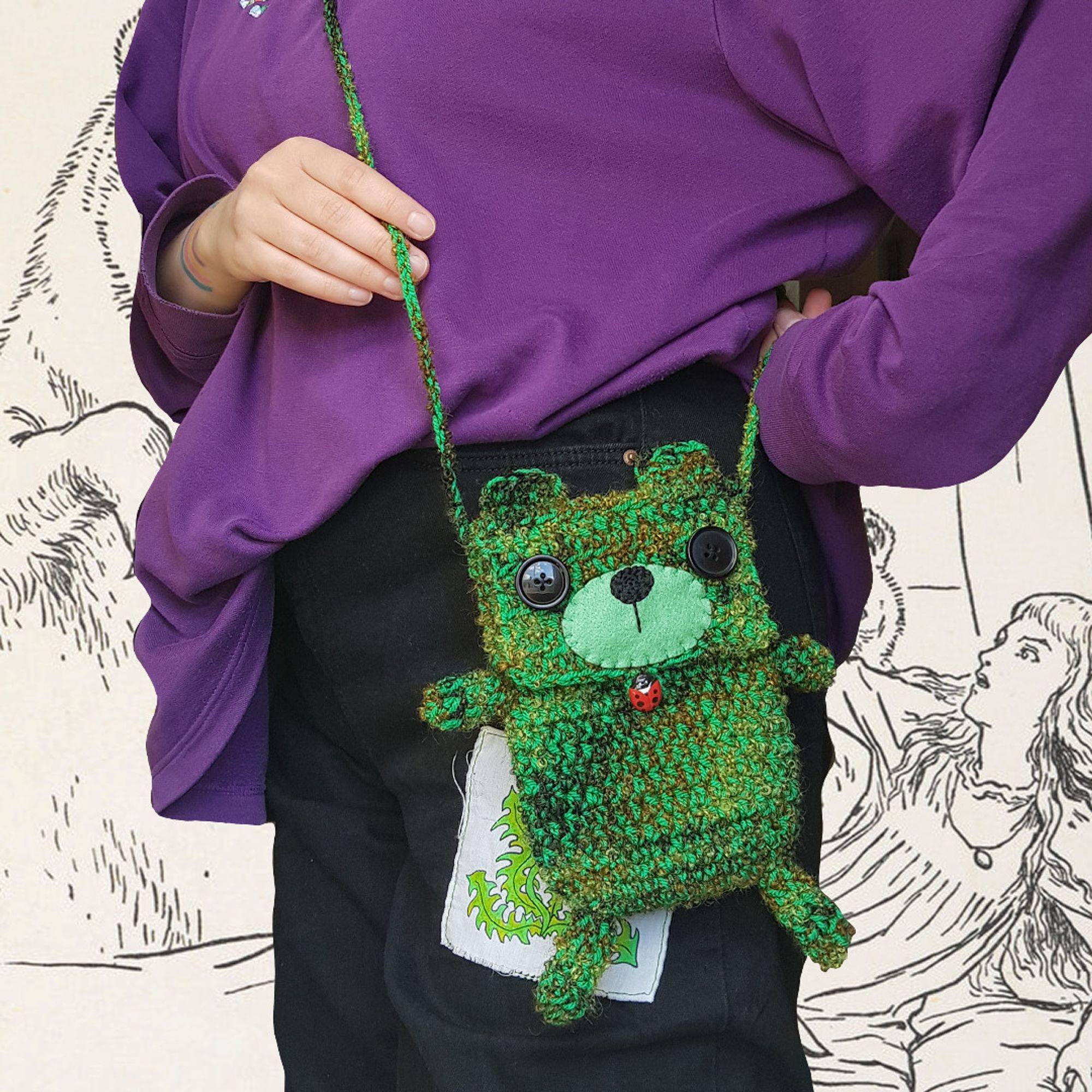 photo of a person side on cropped above the waist and knees they are showing off a cross body, small, green, crochet bag that is shaped like a bear with ears arms and legs, button eyes, felt snout and ladybird button closure.