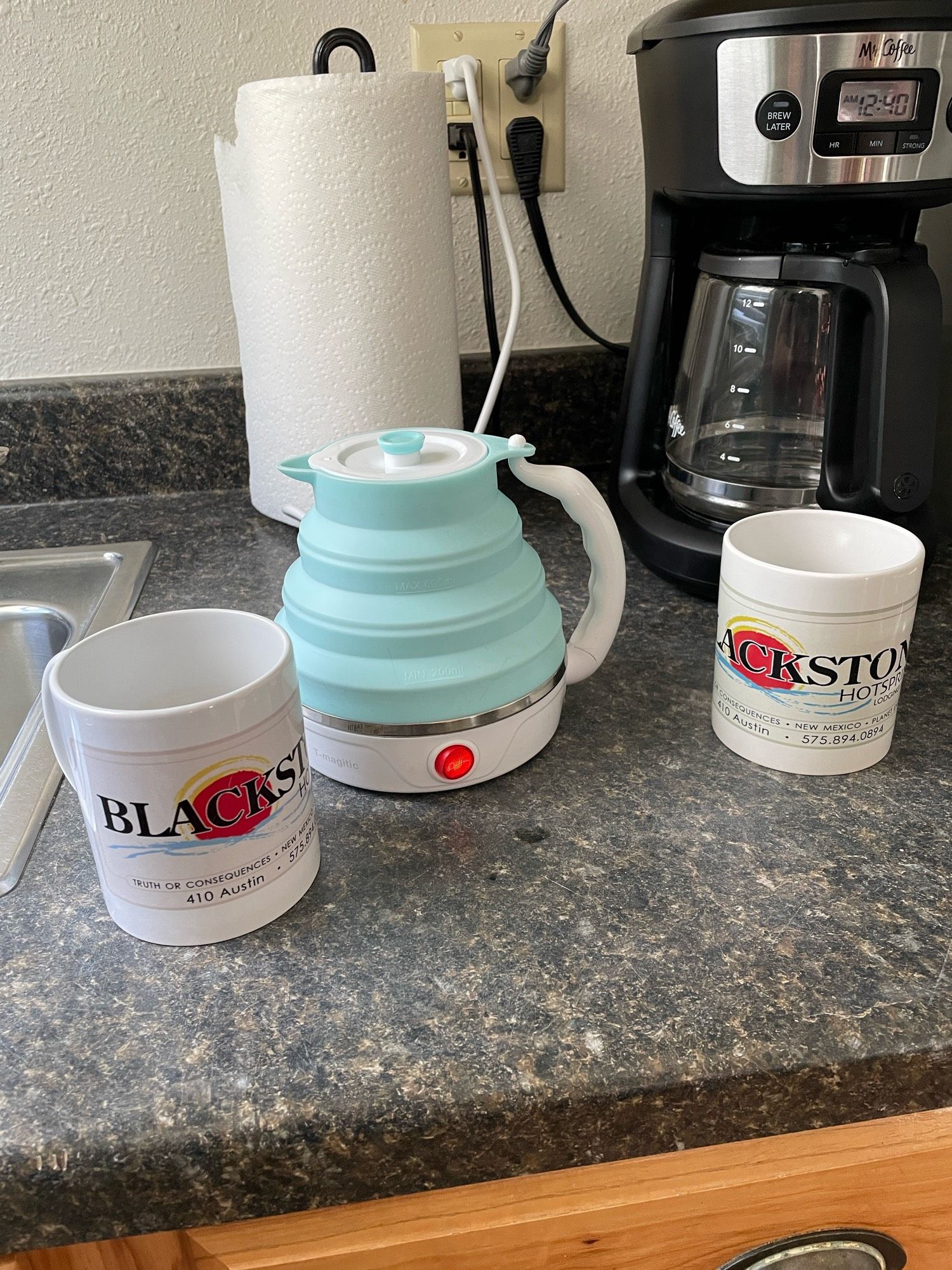 Small silicone electric kettle next to mugs saying Blackstone Hot Springs, sitting in front paper towels and a coffee maker on a dark counter top