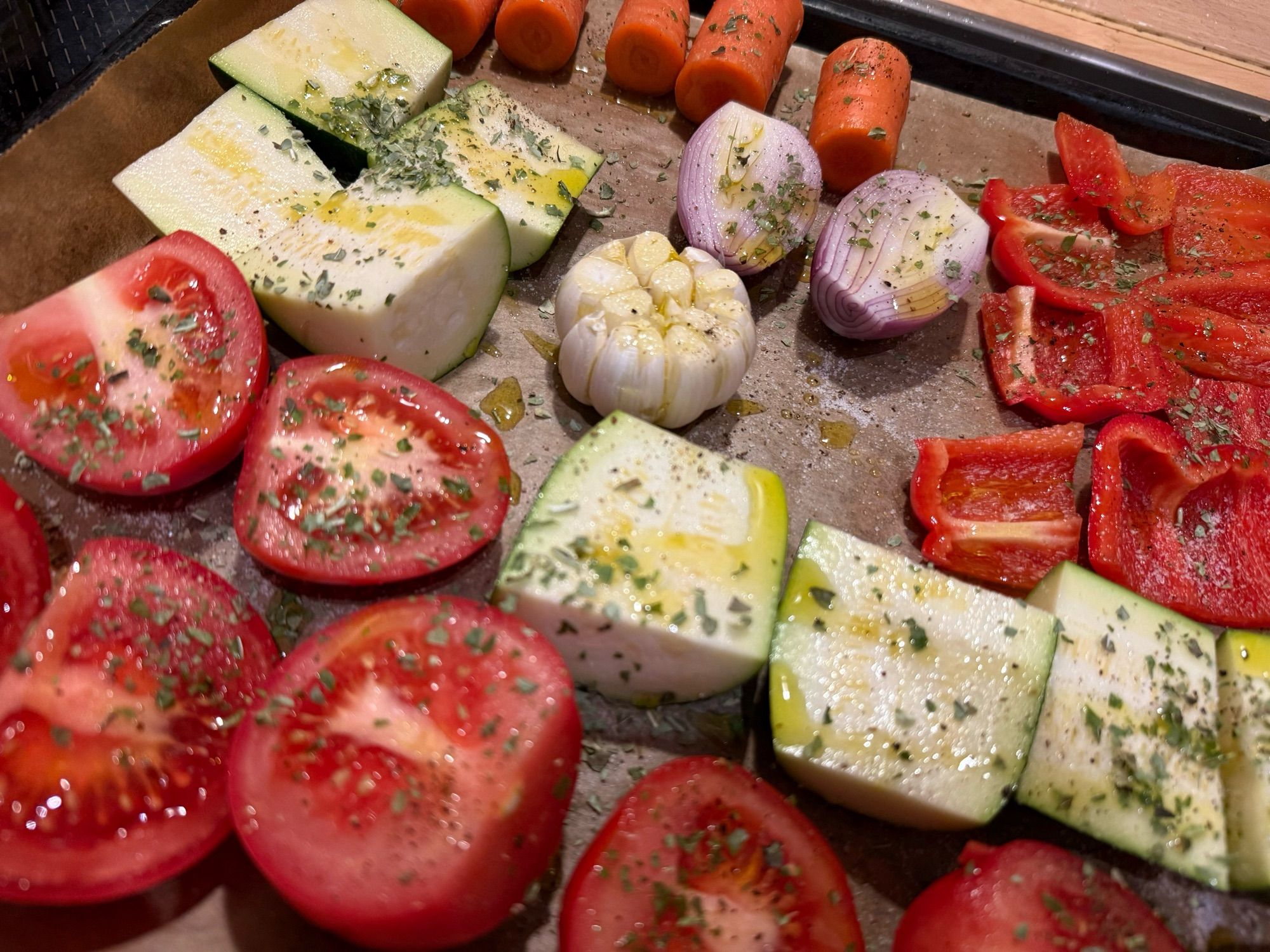 Auf einem Backblech liegen kleingeschnitten und mit Öl und Gewürzen bedeckt: Karotten, rote Paprika, Zucchini, Schalotten, Knoblauch, Tomaten