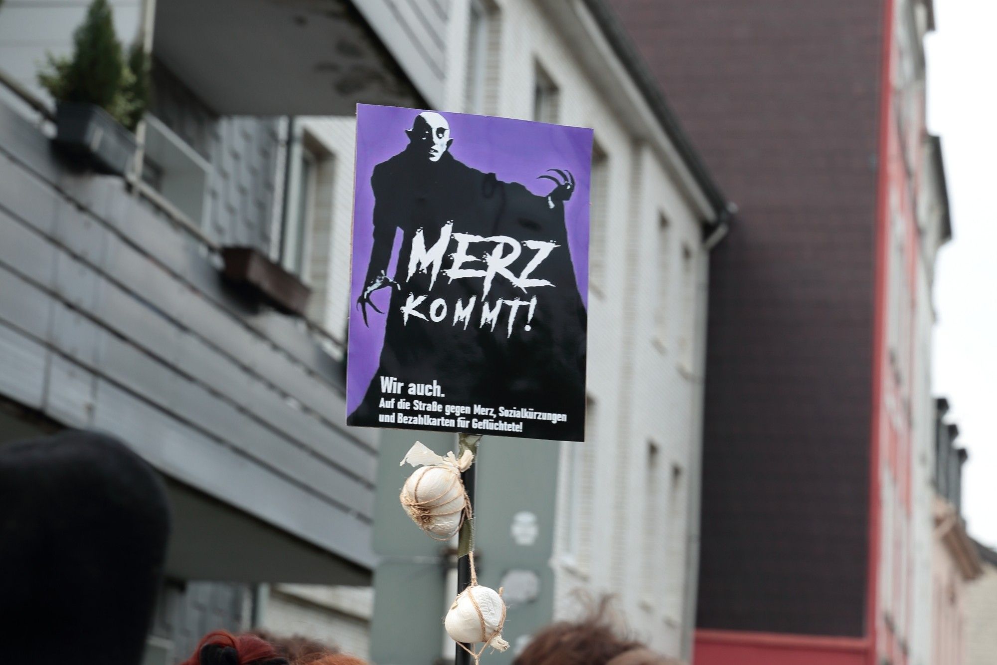 Schild mit einer Nosferatu-Grafik und dem Text "Merz kommt! Wir auch. Auf die Straße gegen Merz, Sozialkürzungen und Bezahlkarten für Geflüchtete!" Am Schilderstiel hängen Knoblauchknollen.