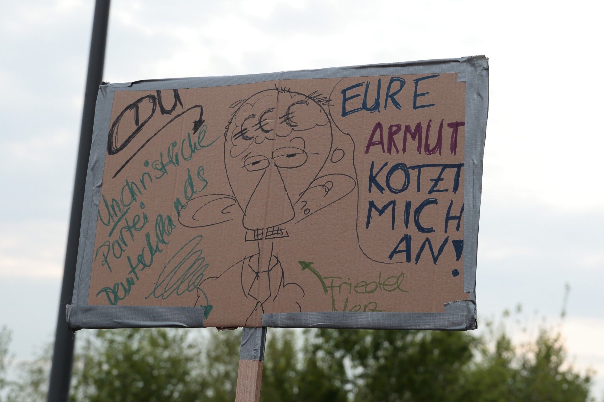 Schild mit Merz Karrikatur und dem Text "CDU Unchristluche Partei Deutschlands. Eure Armut kotzt mich an!"