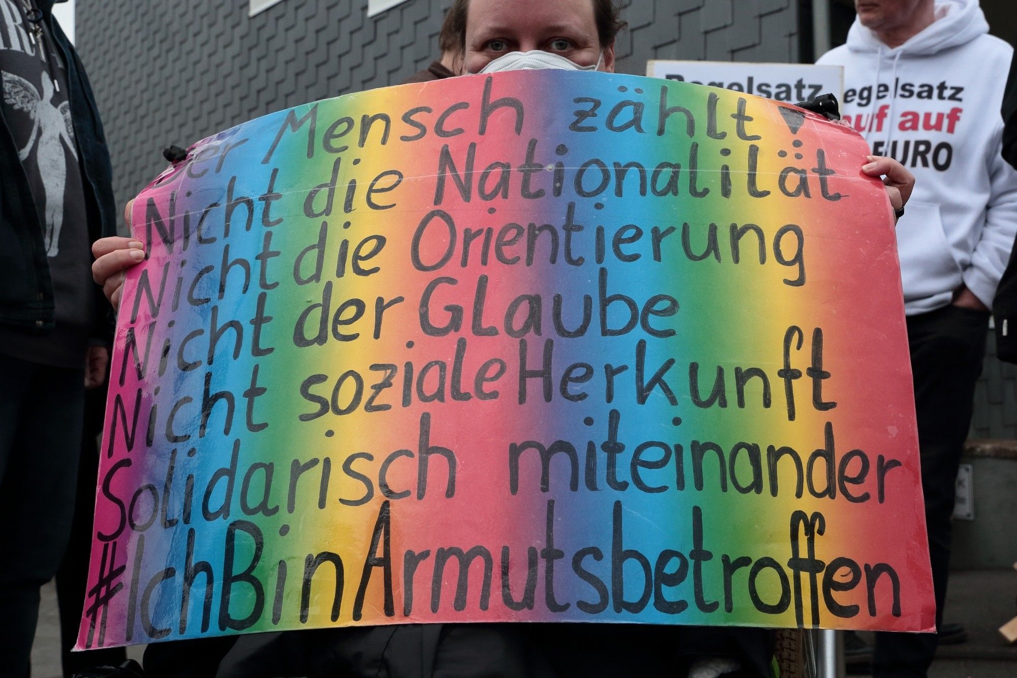 Eine Frau mit FFP2-Maske zeigt ein regenbogenfarbenes Schild mit der Aufschrift "Der Mensch zählt! Nicht die Nationalität. Nicht die Orientierung. Nicht der Glaube. Nicht soziale Herkunft.  Solidarisch miteinander. #IchBinArmutsbetroffen