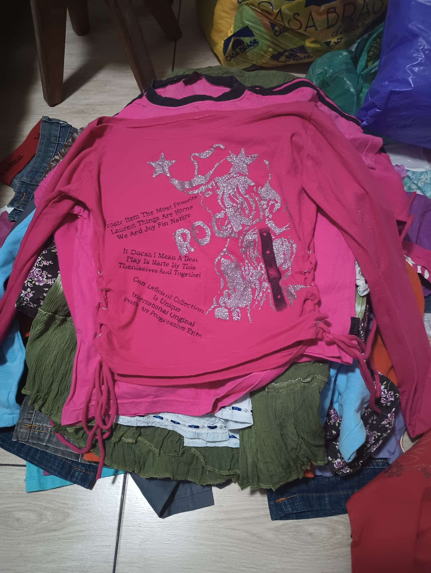 Pilha de roupas de diversas cores no chão. A roupa que está no topo da pilha é uma blusa de mangas longas da cor rosa choque com algumas escritas em inglês, e do lado das escritas há uma mulher de glitter. As roupas são todas dos anos 2000.