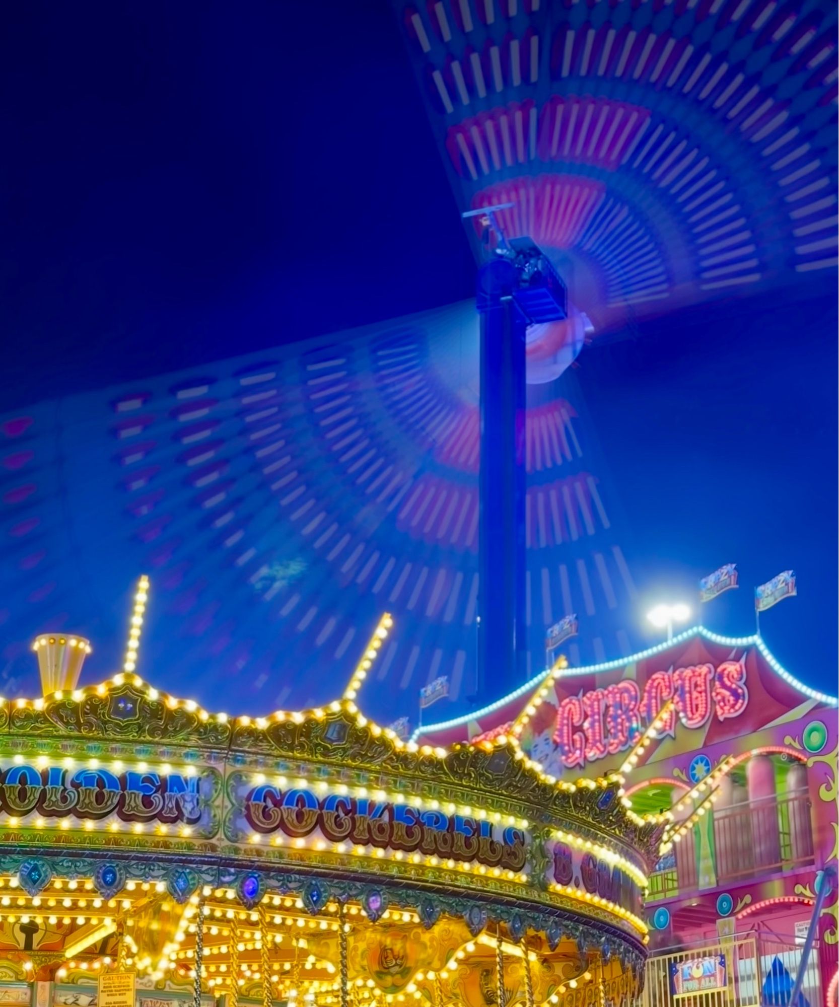 The top of a brightly illuminated carousel in the foreground, with a long exposure in the background showing a ride moving through the air