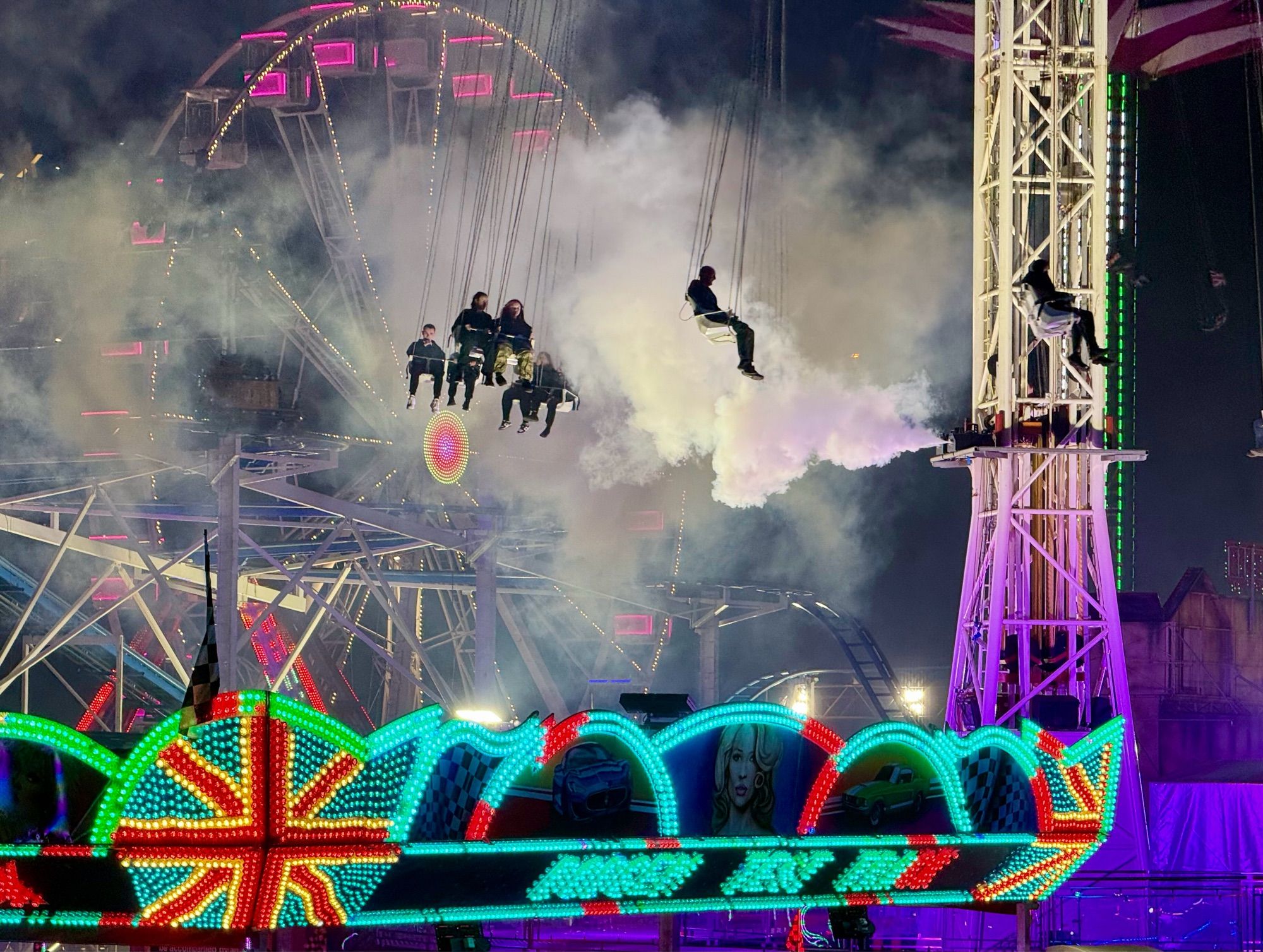 A fun fair with silhouetted people hanging in seats twirling round in the air in front of smoke, with neon lights in the foreground