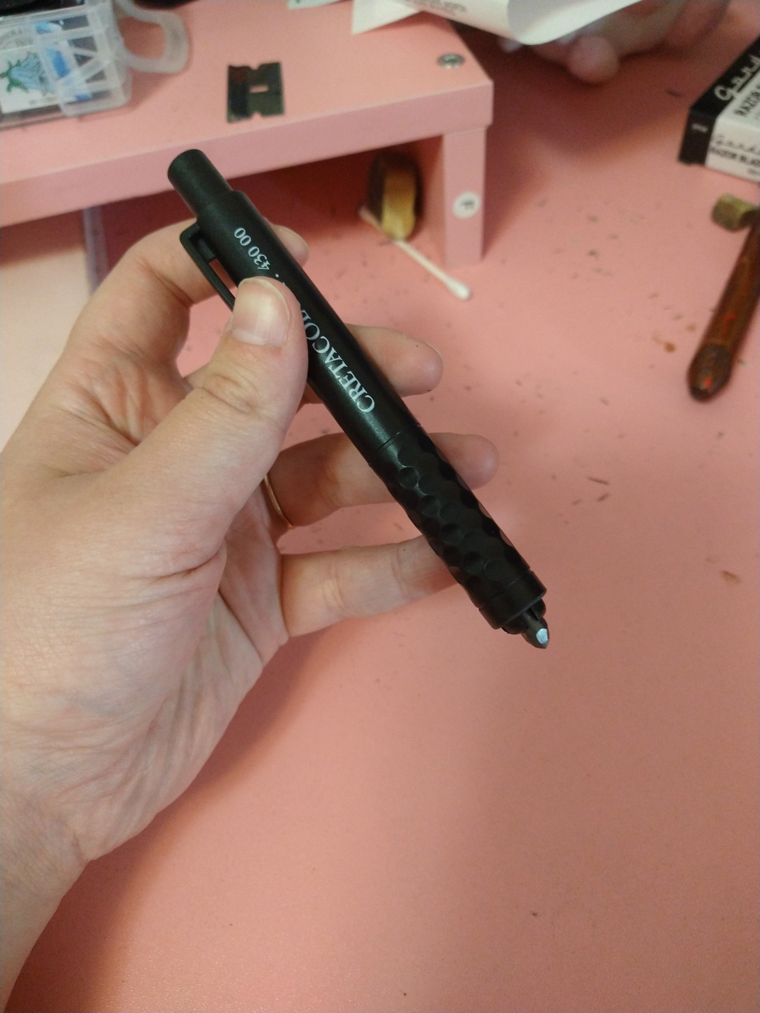 A pale hand holding a 5.6mm black lead holder filled with 2B lead above a pink desk.