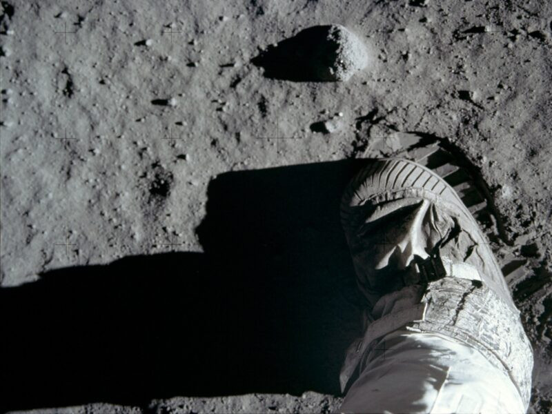 Grey lunar surface with Neil Armstrong's boot lifting up showing the bootprint left behind, wth horizontal strips.