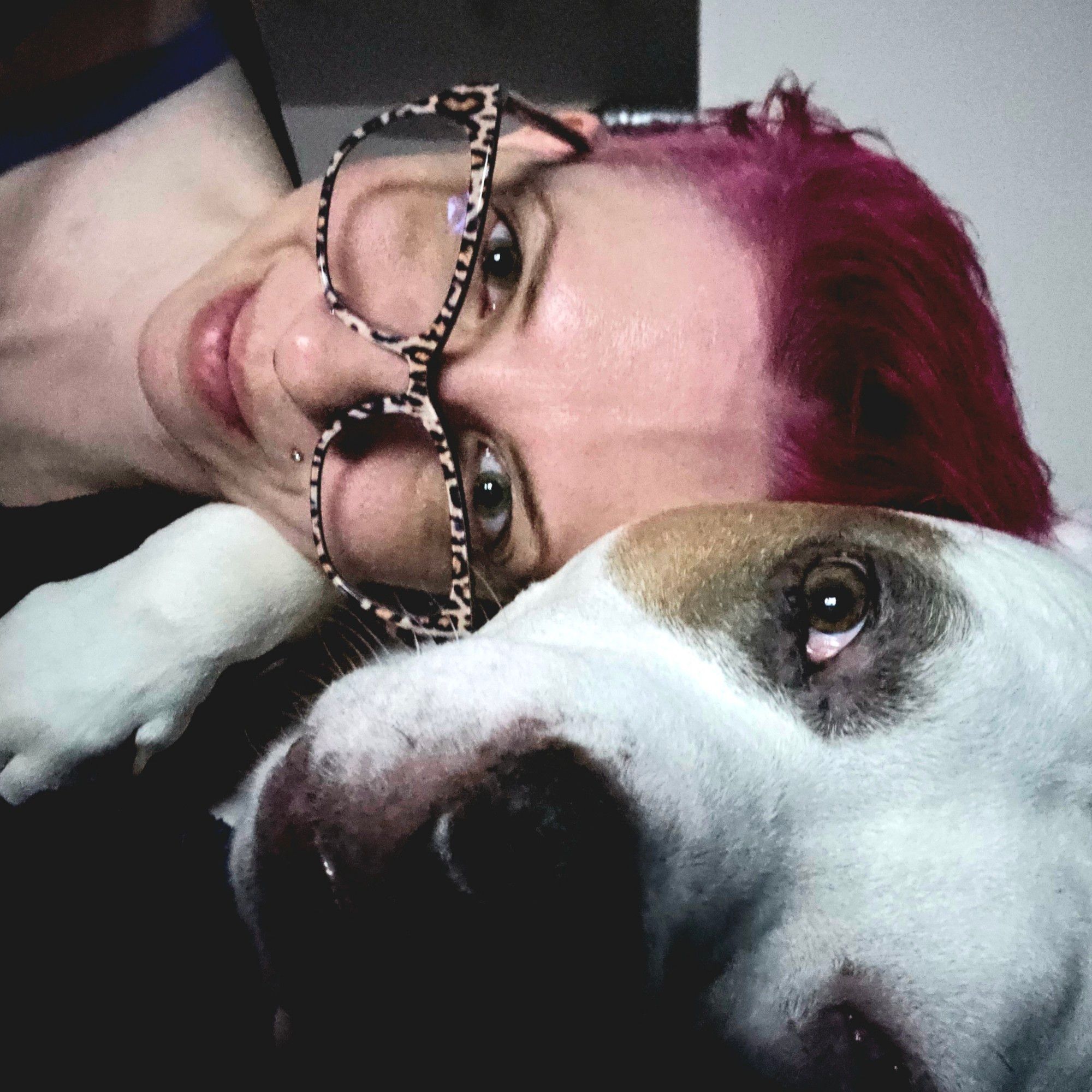 Picture. Kace (femme, white, short dark pink hair, leopard print glasses) uses frankenpup (white pitbull mix brown patches, brown eyes) as a pillow.