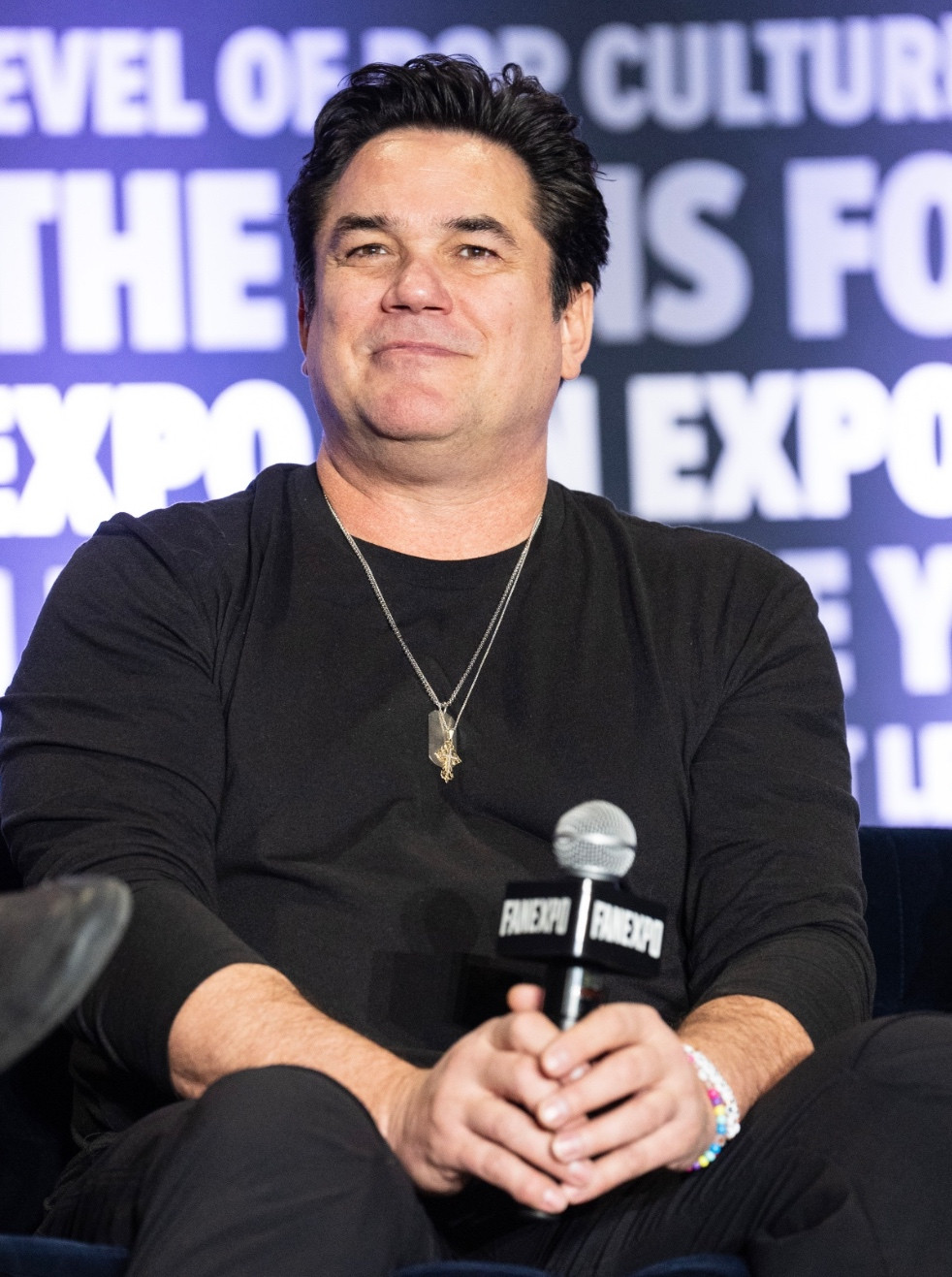 Current photo of Dean Cain, sitting in a chair on a stage, holding a microphone between his sausage fingers. He is wearing all black, which does not hide his obesity. He has the smirk of a mediocre white man above the watery jowls and multiple chins sagging below his skull.