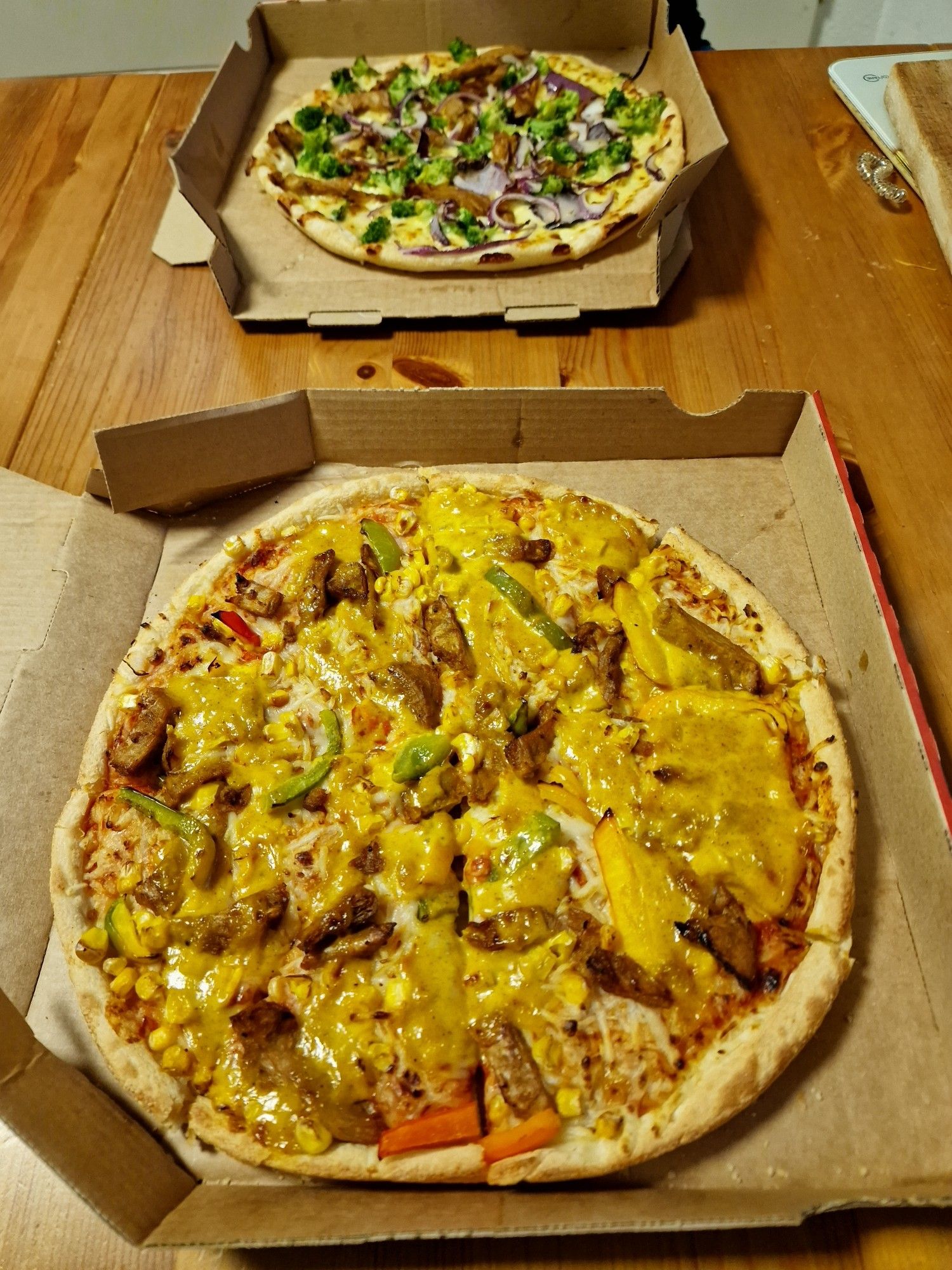 Im Vordergrund ist eine Pizza im Karton mit veganen Filetstücken, Paprika, Mais und Currysauce.
Im Hintergrund ist noch eine Pizza mit Brokkoli.