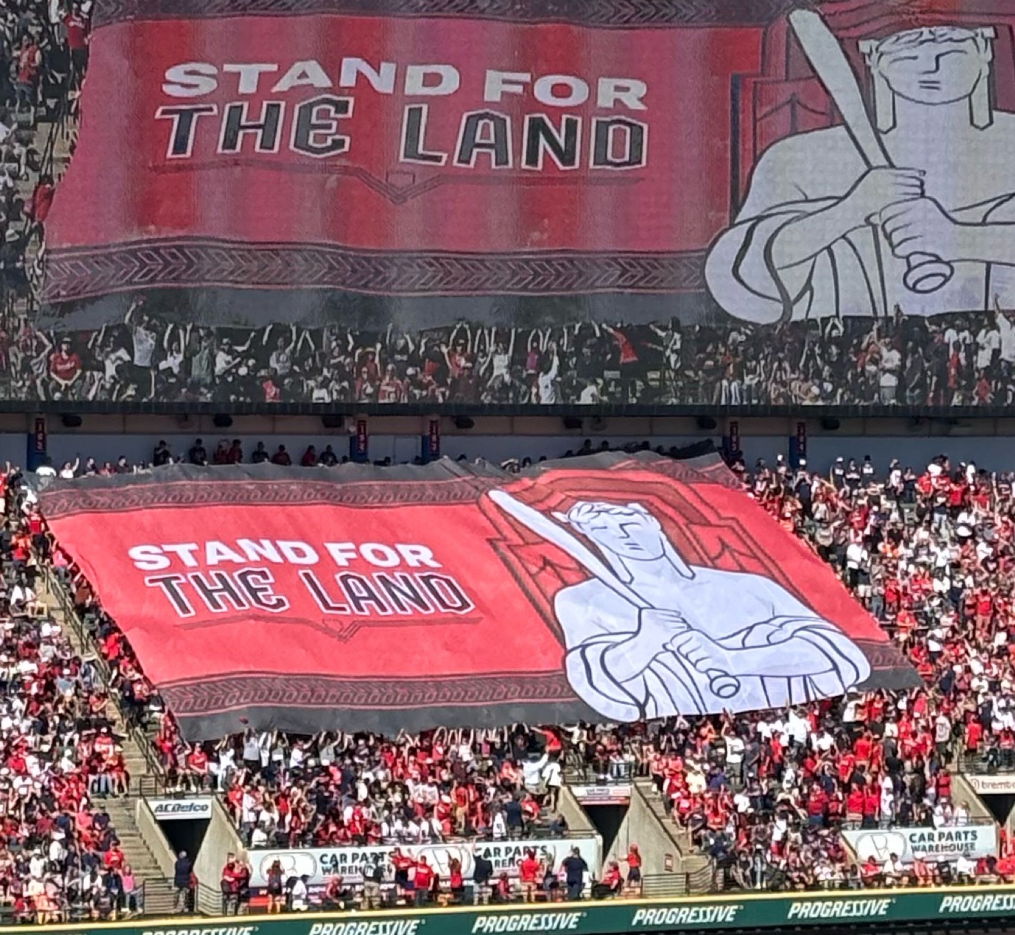 Fans in the bleachers unfurl a giant red banner with a white Cleveland Guardian statue holding a base ball bat and the slogan “Stand for the Land.” The massive video scoreboard above the bleachers shows another angle of this banner.