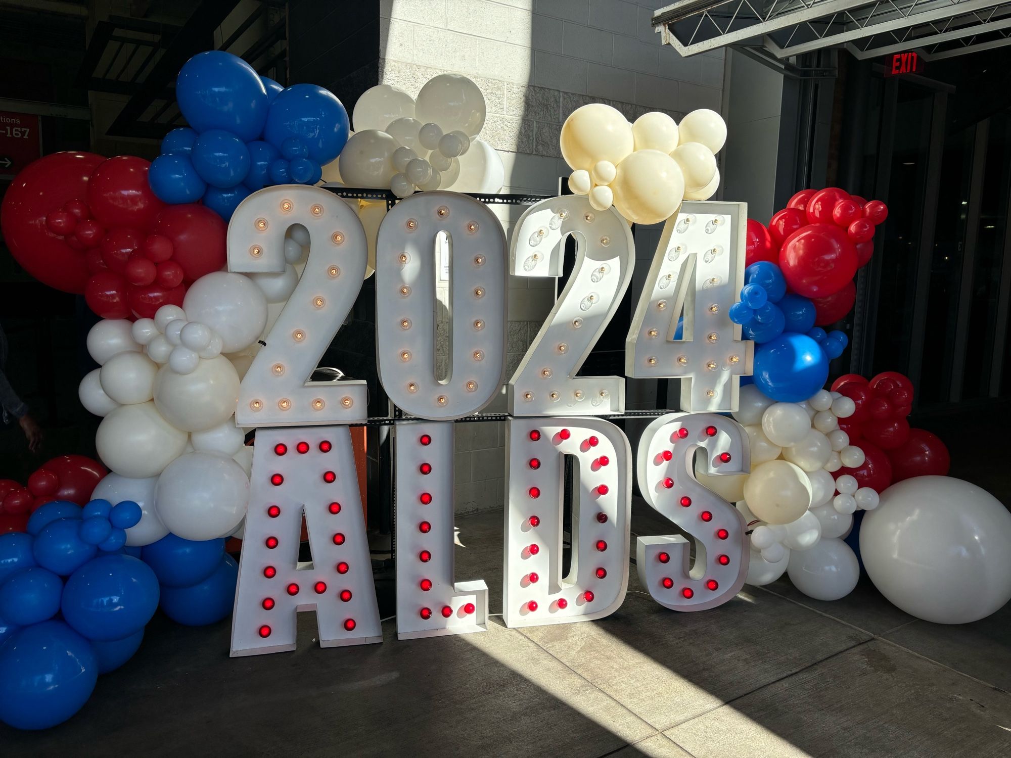 White numbers with white lights atop white letters with red lights reading “2024 ALDS” garnished with red, white, and blue balloons.