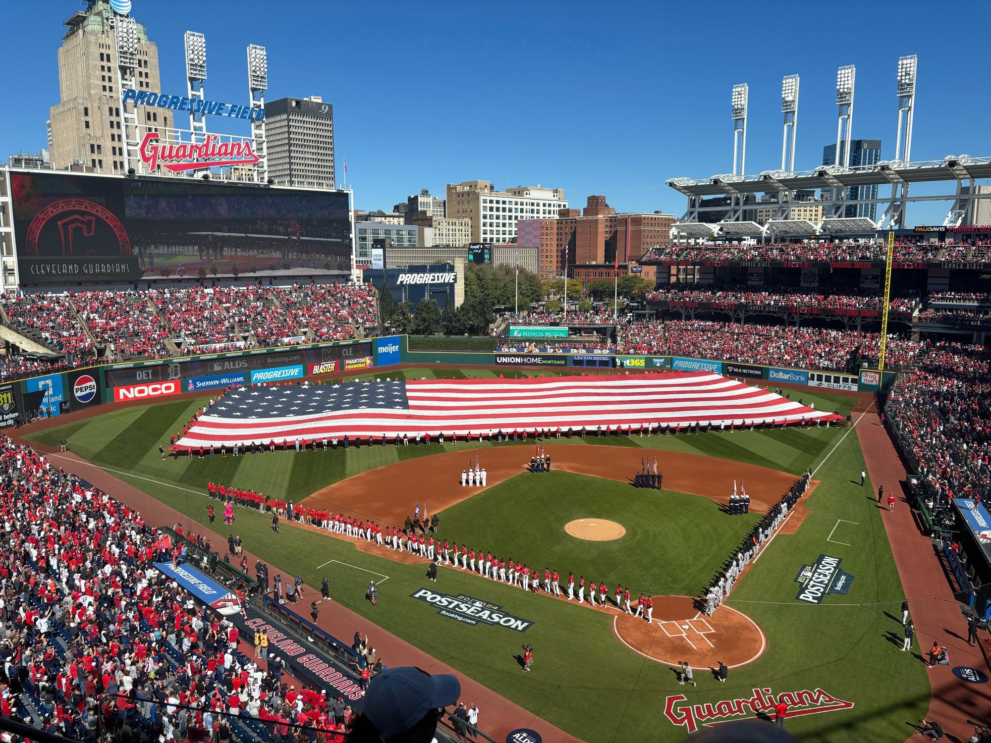 Photo from the upper deck at the Cleveland base ball stadium of the baseball diamond with the Guardians in their red jerseys and white pants along the third base line, Tigers in their road grays along the first, with a giant USA flag being carried over the outfield.