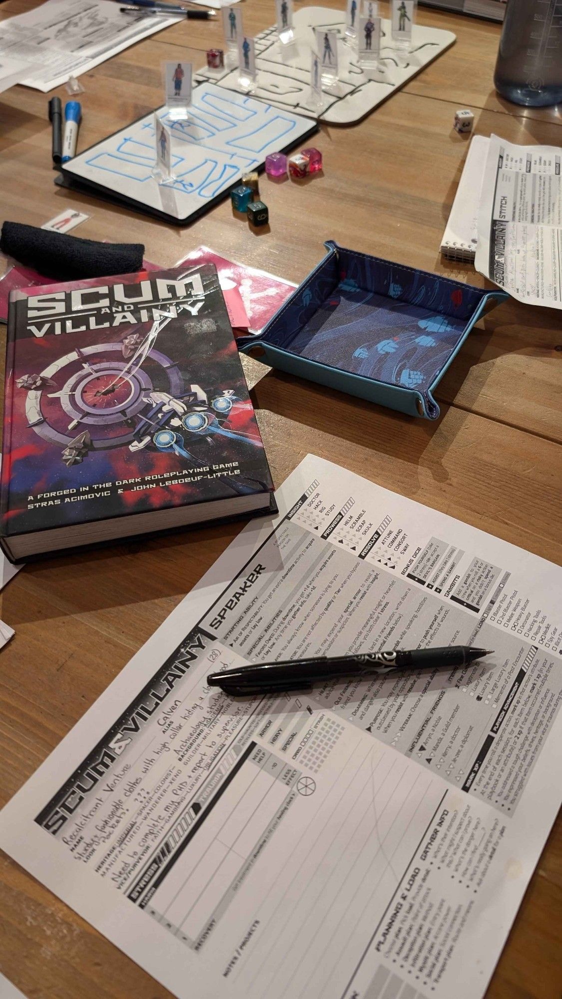 The Scum and Villainy rulebook, dice and a dice tray, a character sheet and some paper standees for characters laid out on a table.