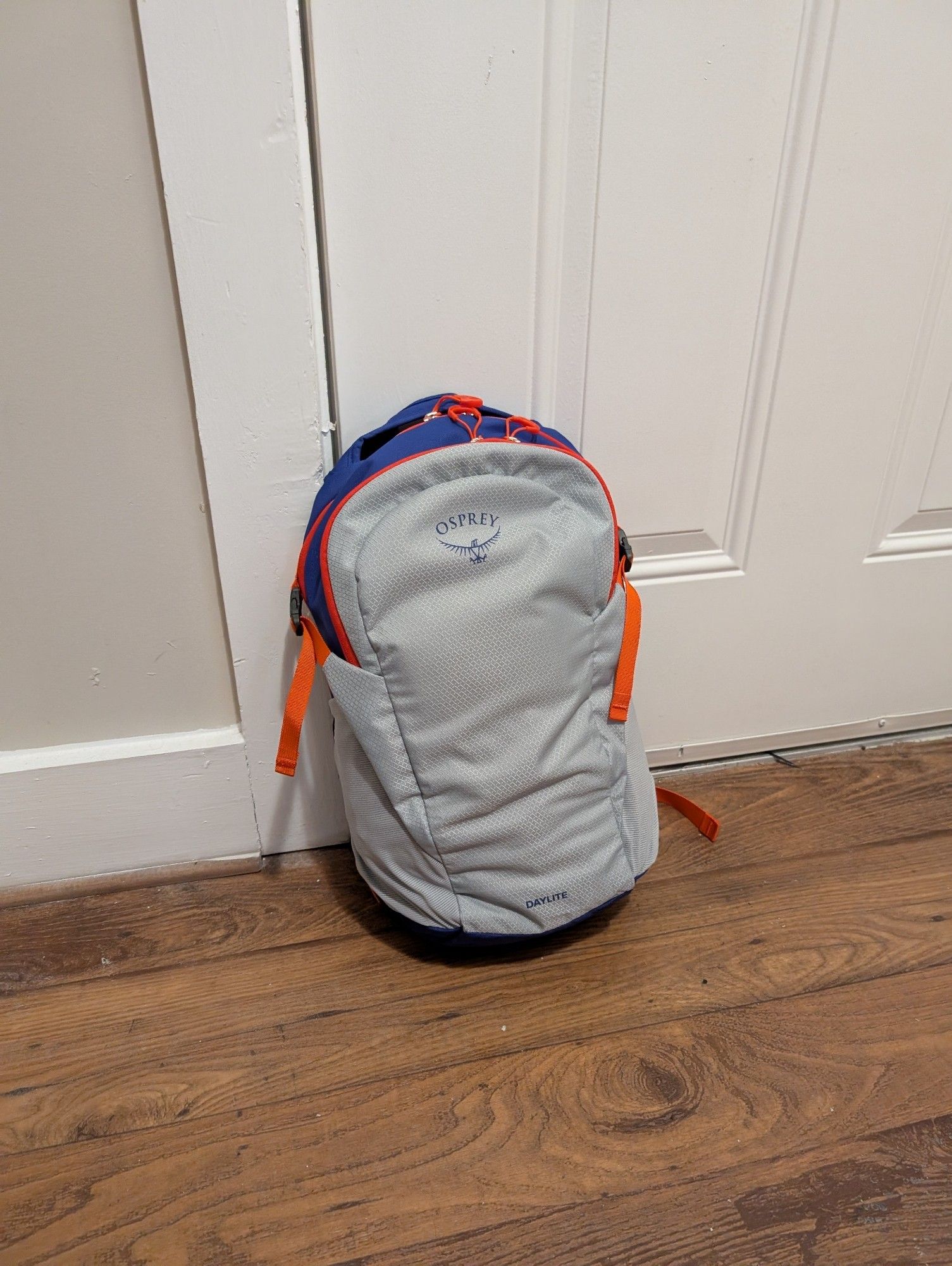 Small blue, orange and gray Osprey daypack, not quite completely stuffed, leaning against a door