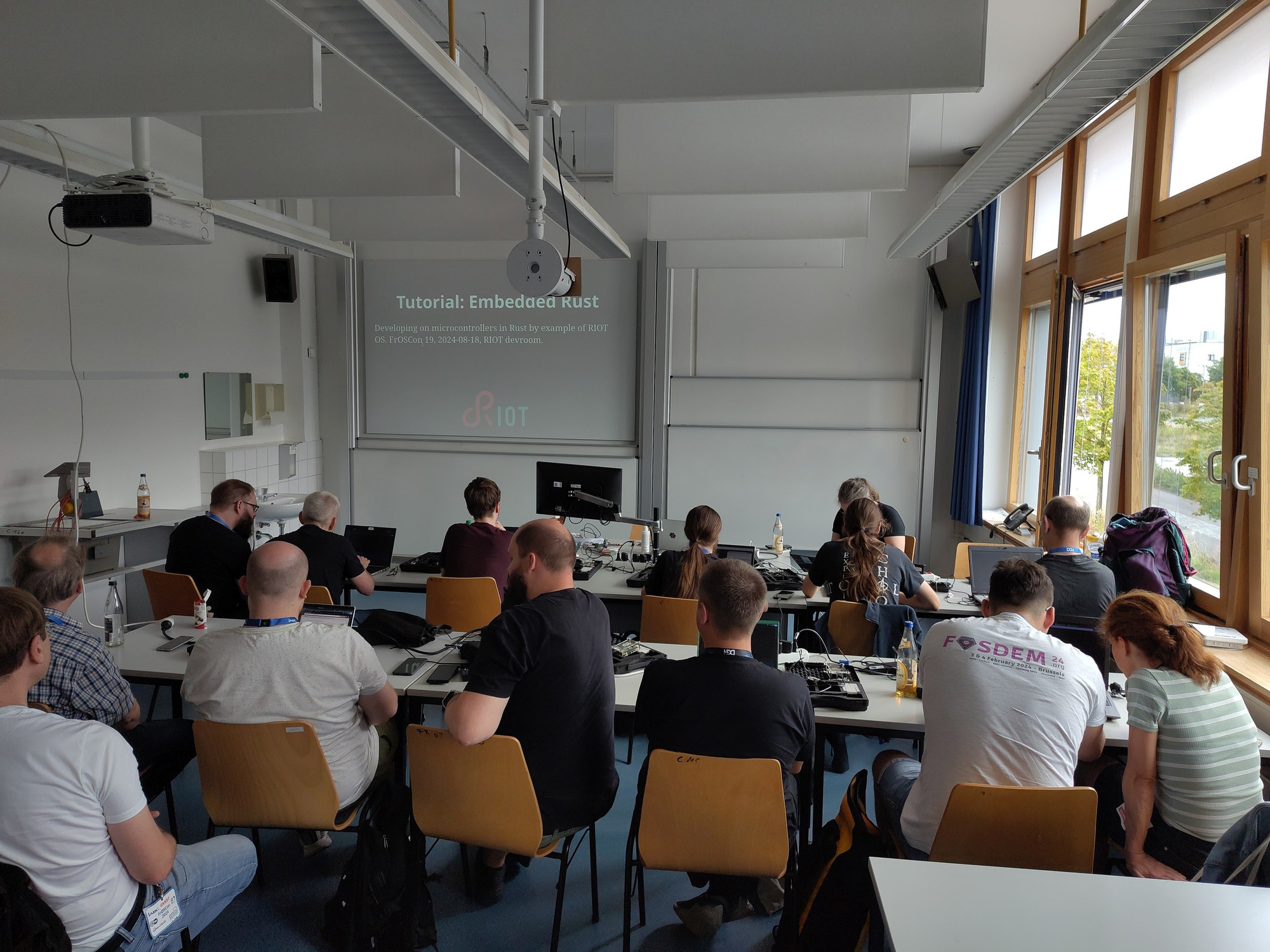 A small auditorium with over a dozen people working in small groups on laptops and hardware.

A slide says "Tutorial: Embedded Rust".