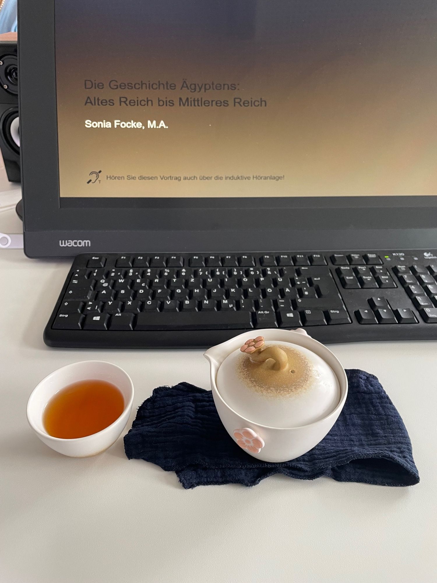 Travel tea set in white with a light-brown dip at the bottom. The teapot, sitting on a blue tea towel, has pink flowers on either side for the fingers and another pink flower on top. The cup is filled with a reddish herbal tea. Behind them, a black keyboard and a wacom tablet displaying a PowerPoint presentation.