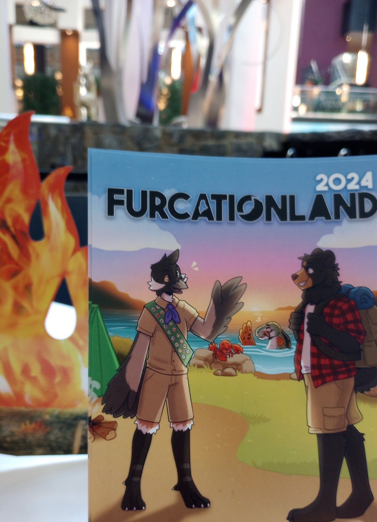 Convention poster with friendly anthro bird and bear. The photo taken, while indoors, is situated right next to a fake campfire that tries to blend with part of a campfire and tent in the poster.