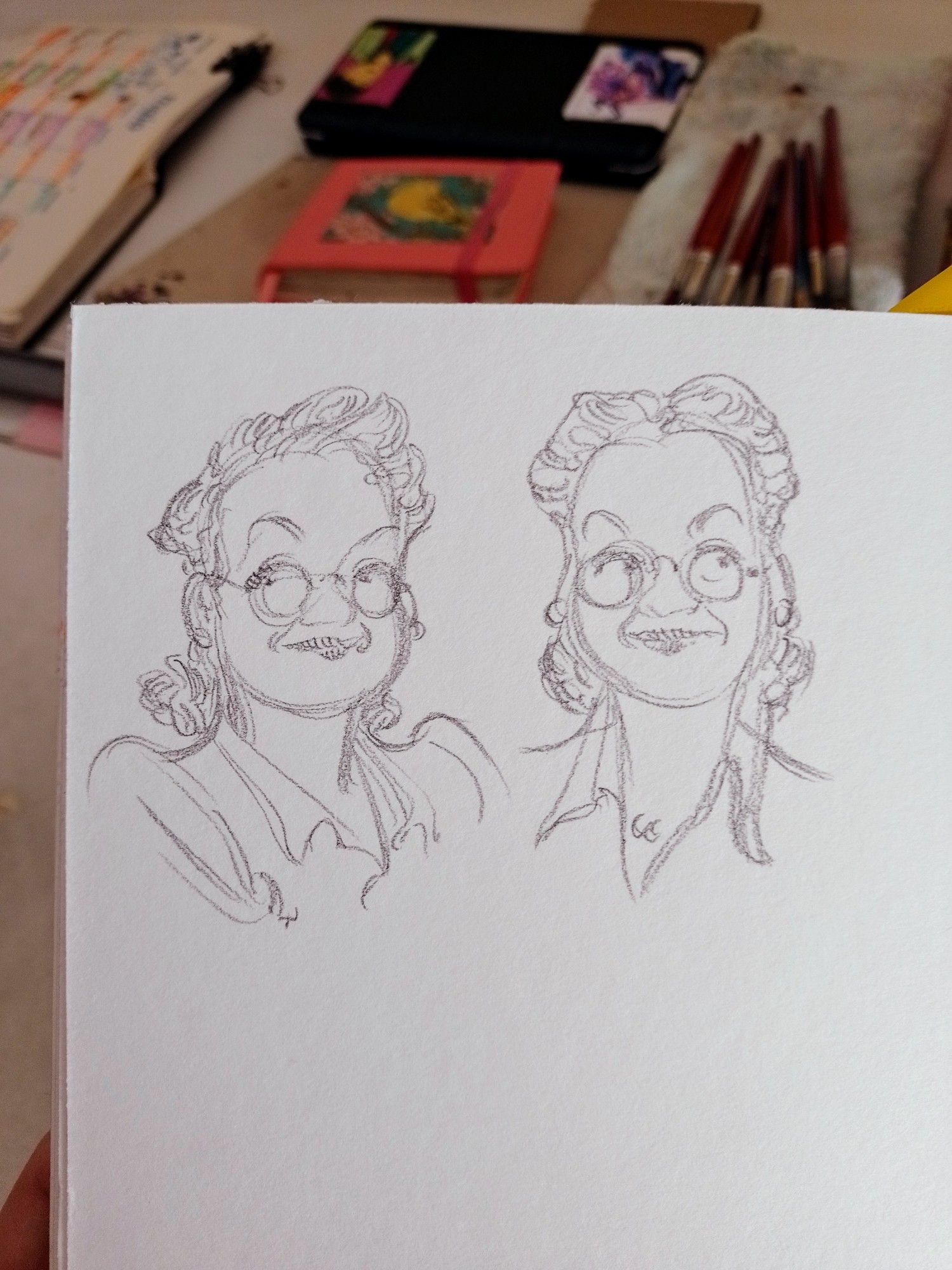 Foto colorida de parte de meu sketchbook mostrando estudos da personagem Vitória mais velha, usando óculos e com penteado estilo anos 1940. Ao fundo, a mesa de desenho com pincéis, sketchbooks e outros materiais.