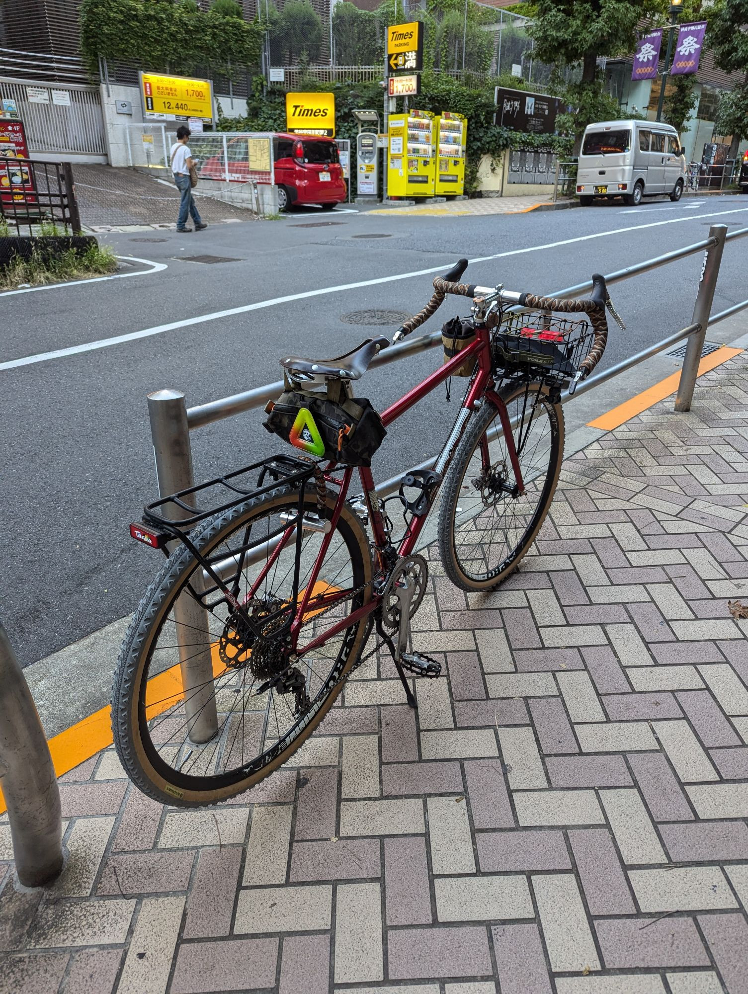 Touring bike with a steel red frame with grey ultradynamico tires, a rear rack, front basket, saddle bag attached to a Brooks saddle. The wide gravel bars have patterned bar tape, and bar end friction shifters.