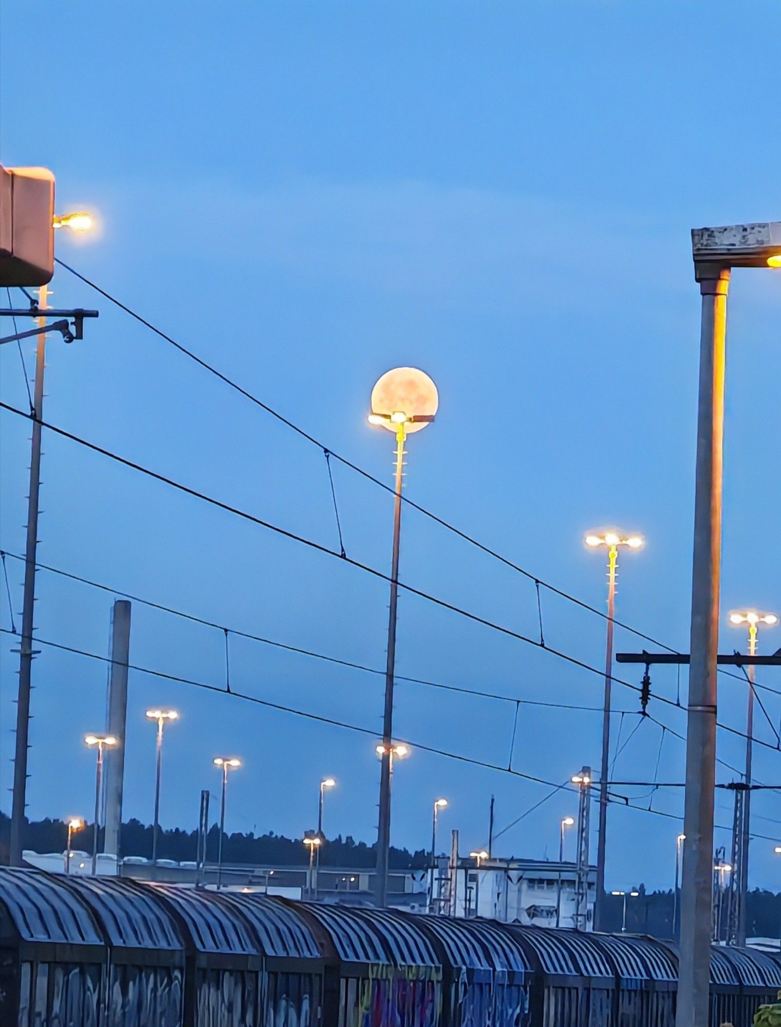 Ein Rangierbahnhof. Am unteren Bildrand Güterwaggons, teils mit Graffiti besprüht. Im Bild verteilen sich Licht- und Oberleitungsmasten, Stromleitungen ziehen sich quer durch das Bild. Am Himmel der orangene Vollmond "gestützt" auf einem Lichtmast. Blauer Himmel kurz vor Sonnenaufgang.