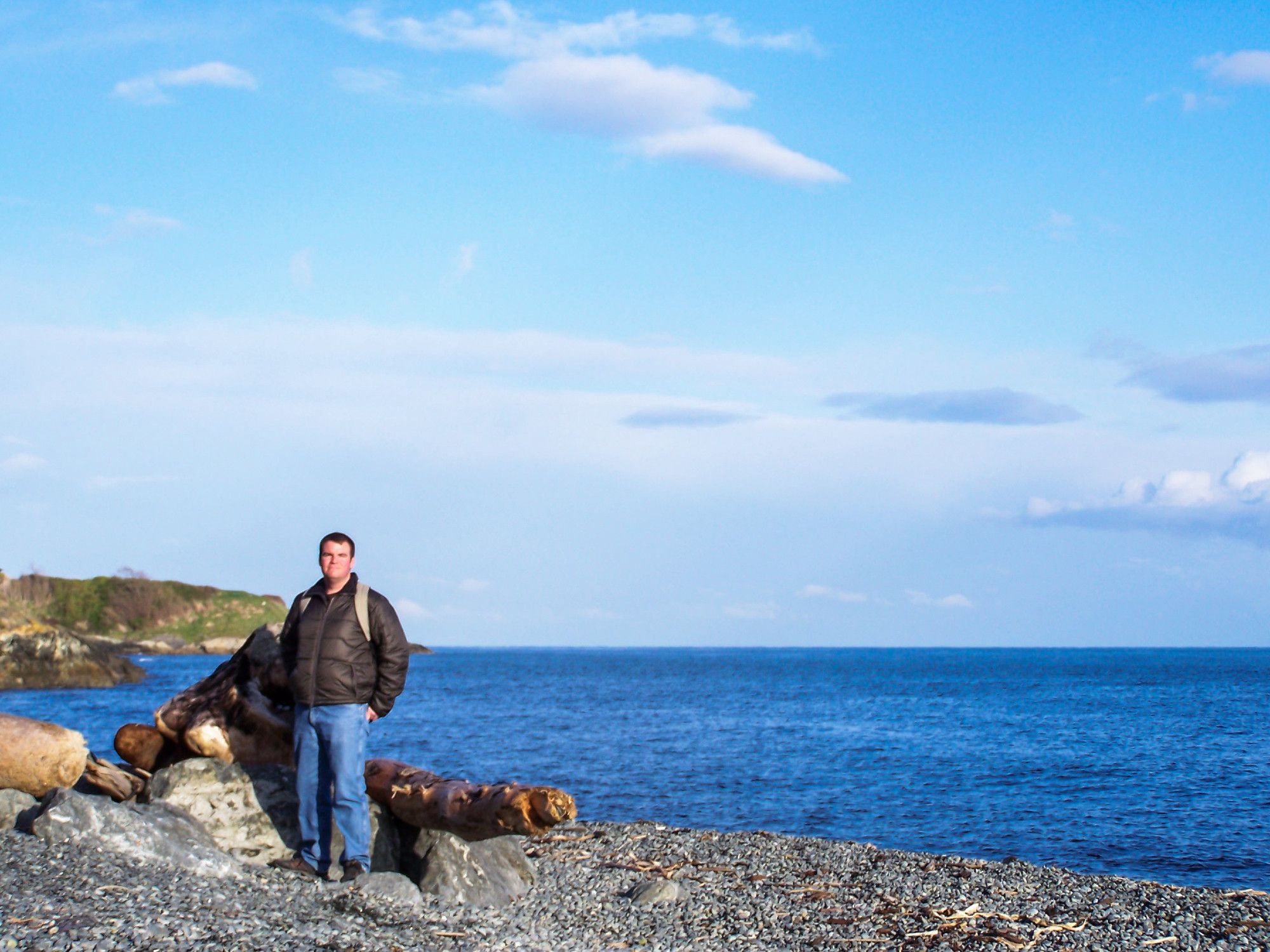 Me standing on a pebbly beach in front of drift logs and the blue ocean.