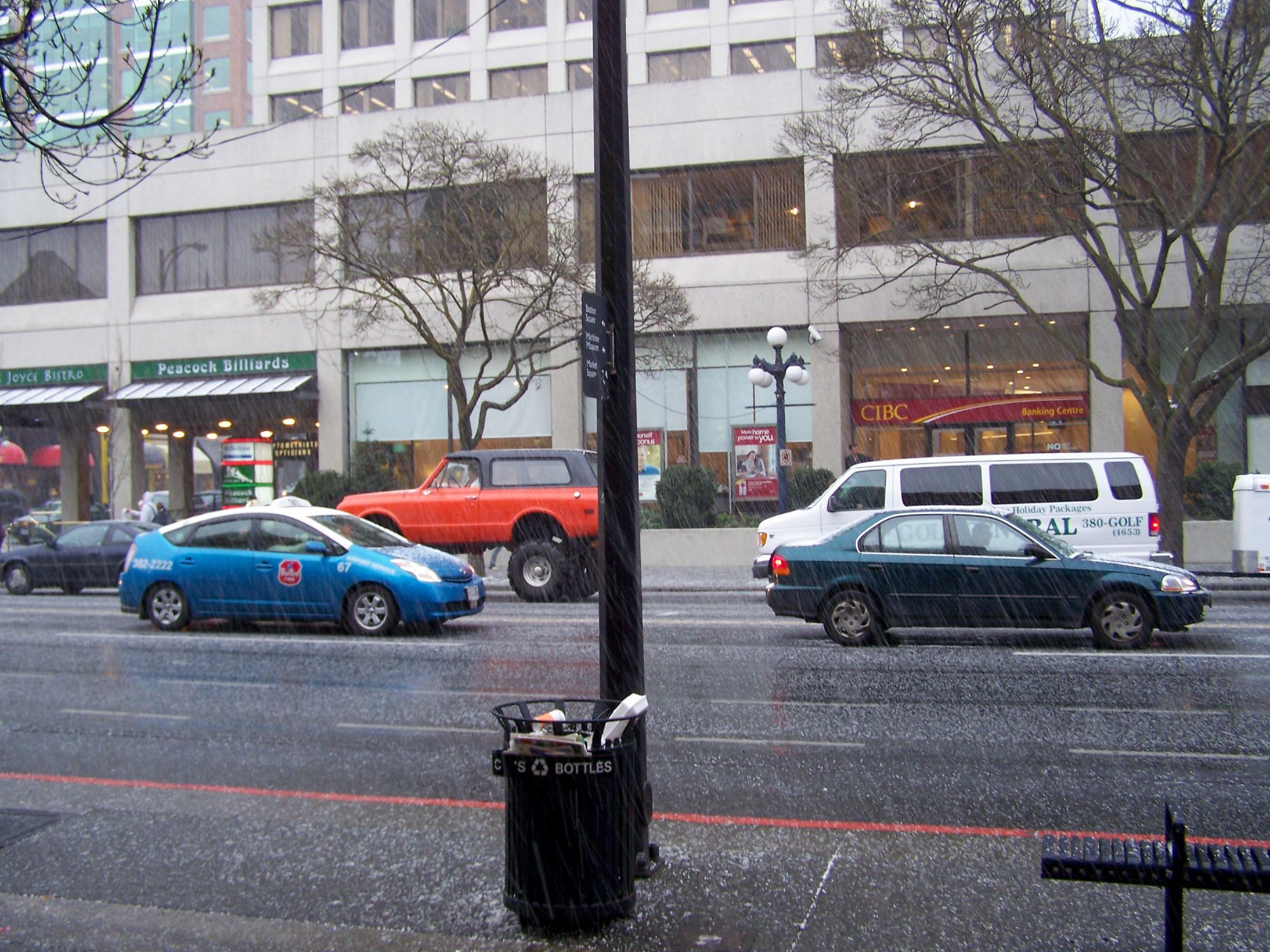 A blue and green car going to the right and orange SUV and white van going to the left on a busy downtown street with a tall whitebuilding in the background and garbage can, light pole and bench  in the foreground