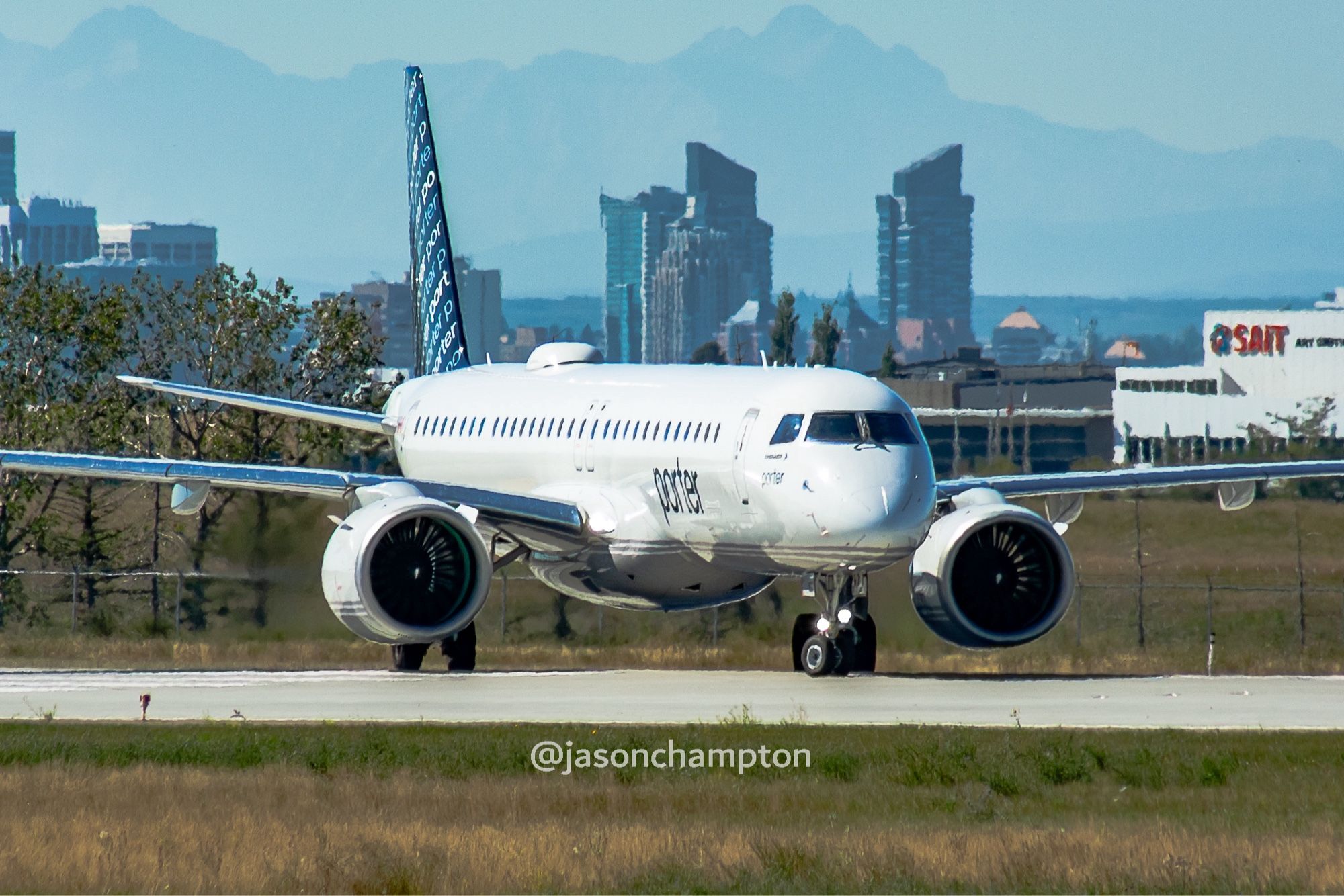 A white jetliner with navy blue tail and Porter wordmark in navy in the fuselage. The City of Calgary skyline and SAIT building in the background