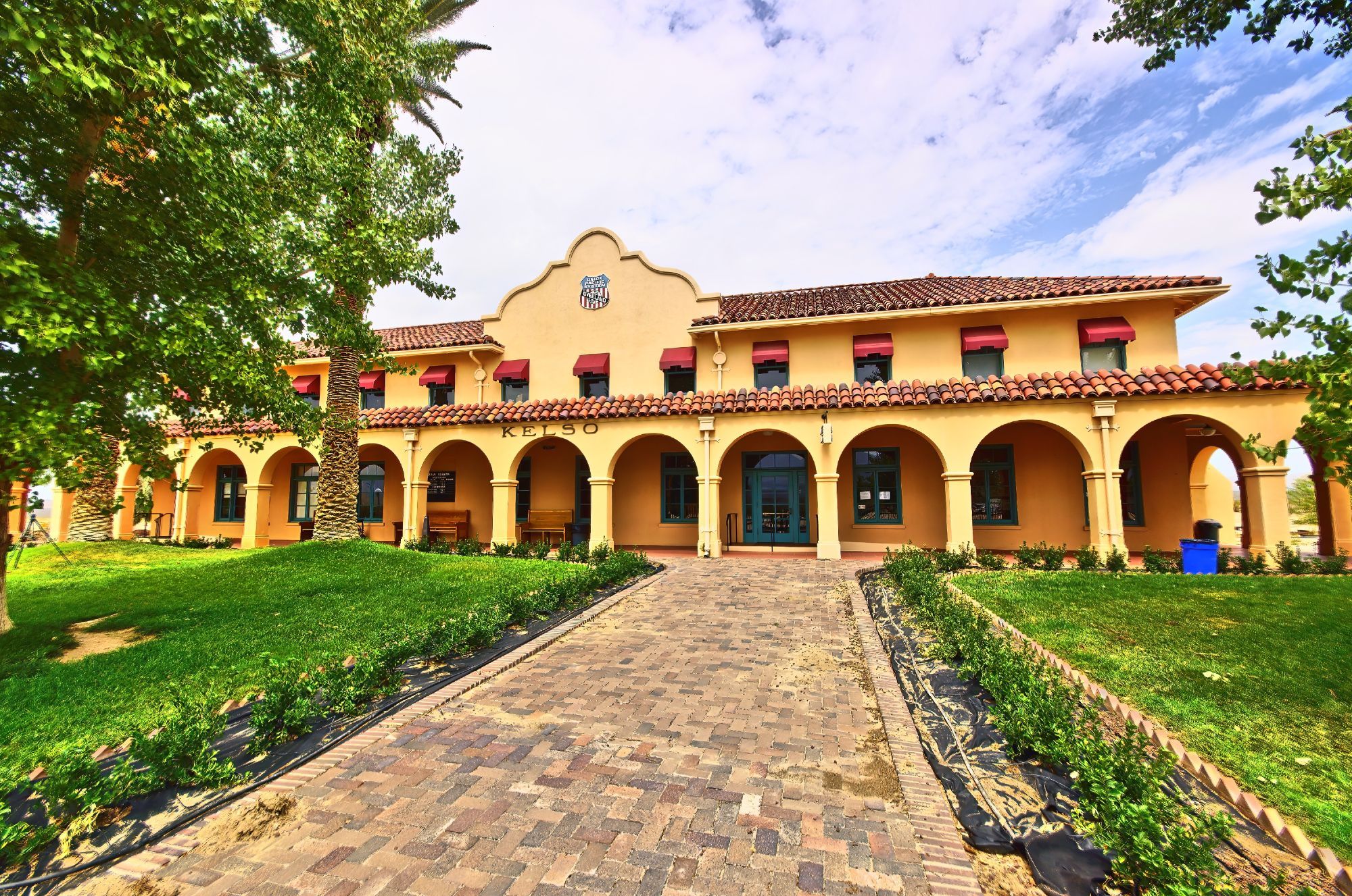 On the left and right, green lawns with trees on the left. Centre is a wide brick walkway lined with small shrubs. The walkway and the lawns lead up to a two story tan coloured building with a red tiled roof. The lower level has a veranda fronted by 11 arches. The architectural style is mission revival